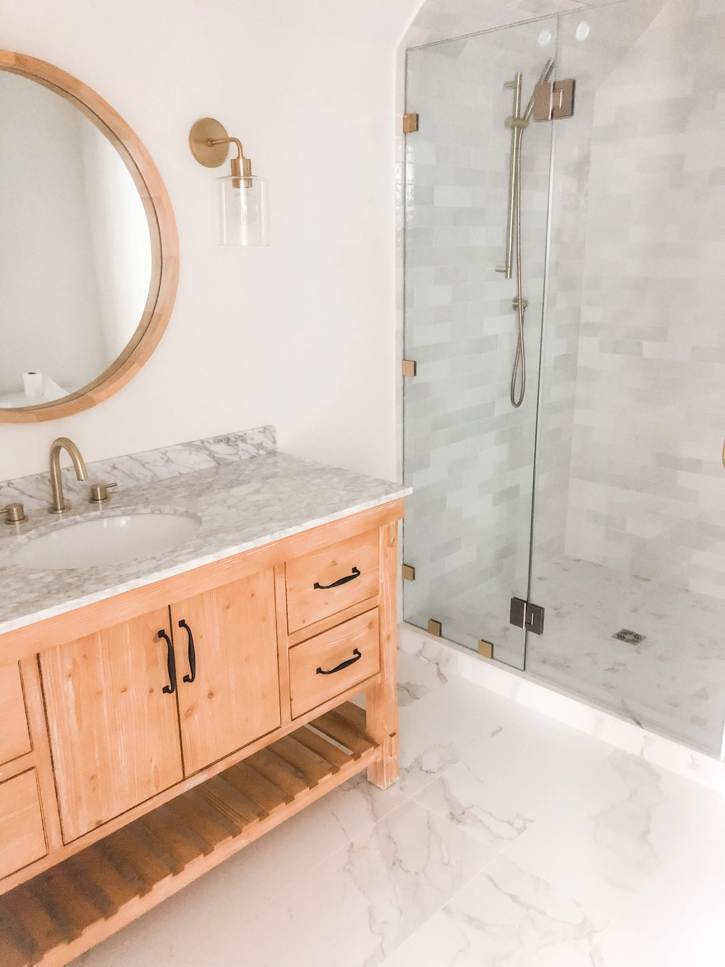 Bathroom with wooden vanity, marble countertop, round mirror, wall light, and glass shower enclosure with watercolor white tiles.