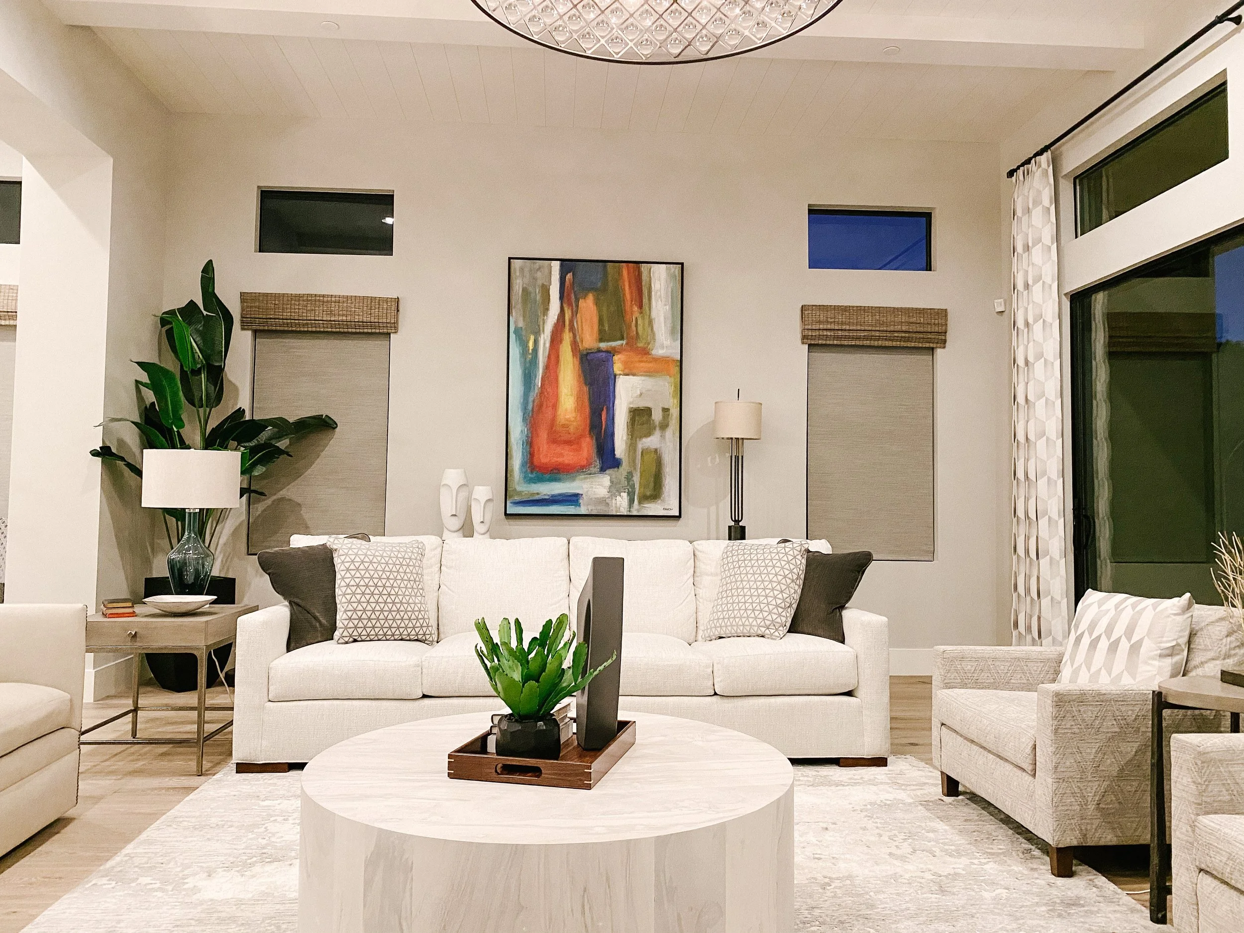 Living room with white sofa, beige armchair, abstract painting, large green plant, floor lamp, coffee table with green plant, and windows with blinds.