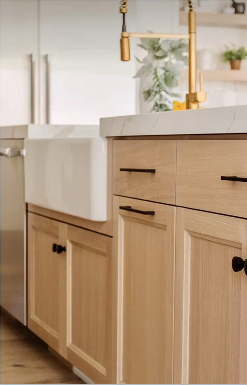 Close-up of a kitchen cabinet made of light wood with black handles, a white marble countertop, a gold faucet, and some plants in the background.