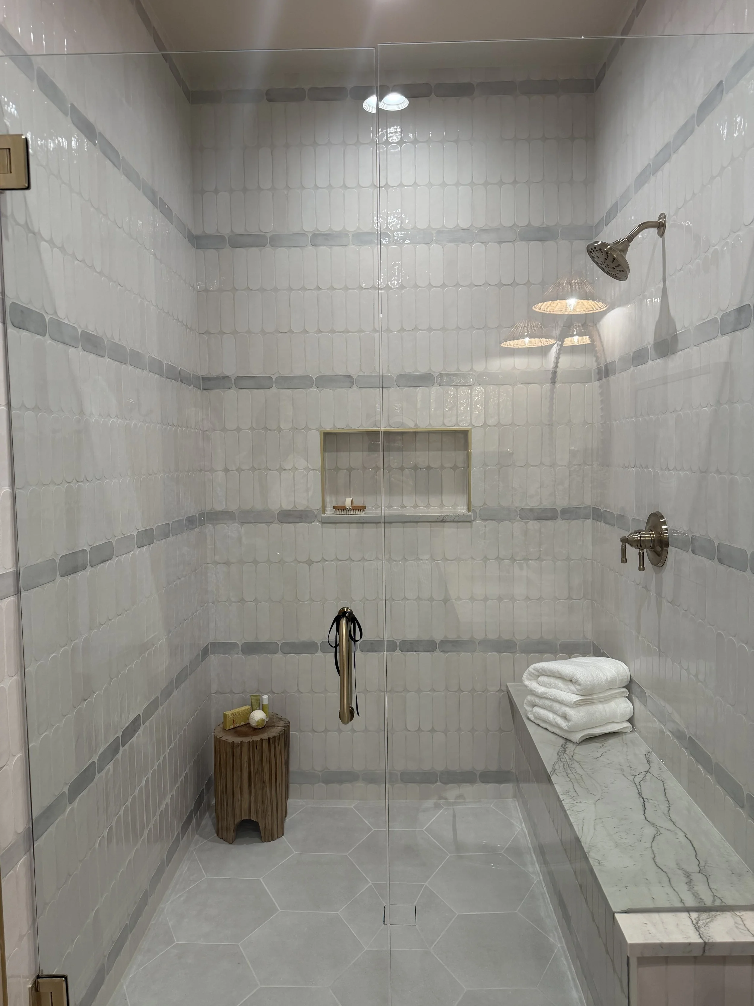 Modern shower with white and gray tiles, glass door, a shower head, a shelf with toiletries, a small wooden stool with toiletries, and a marble bench with folded towels.