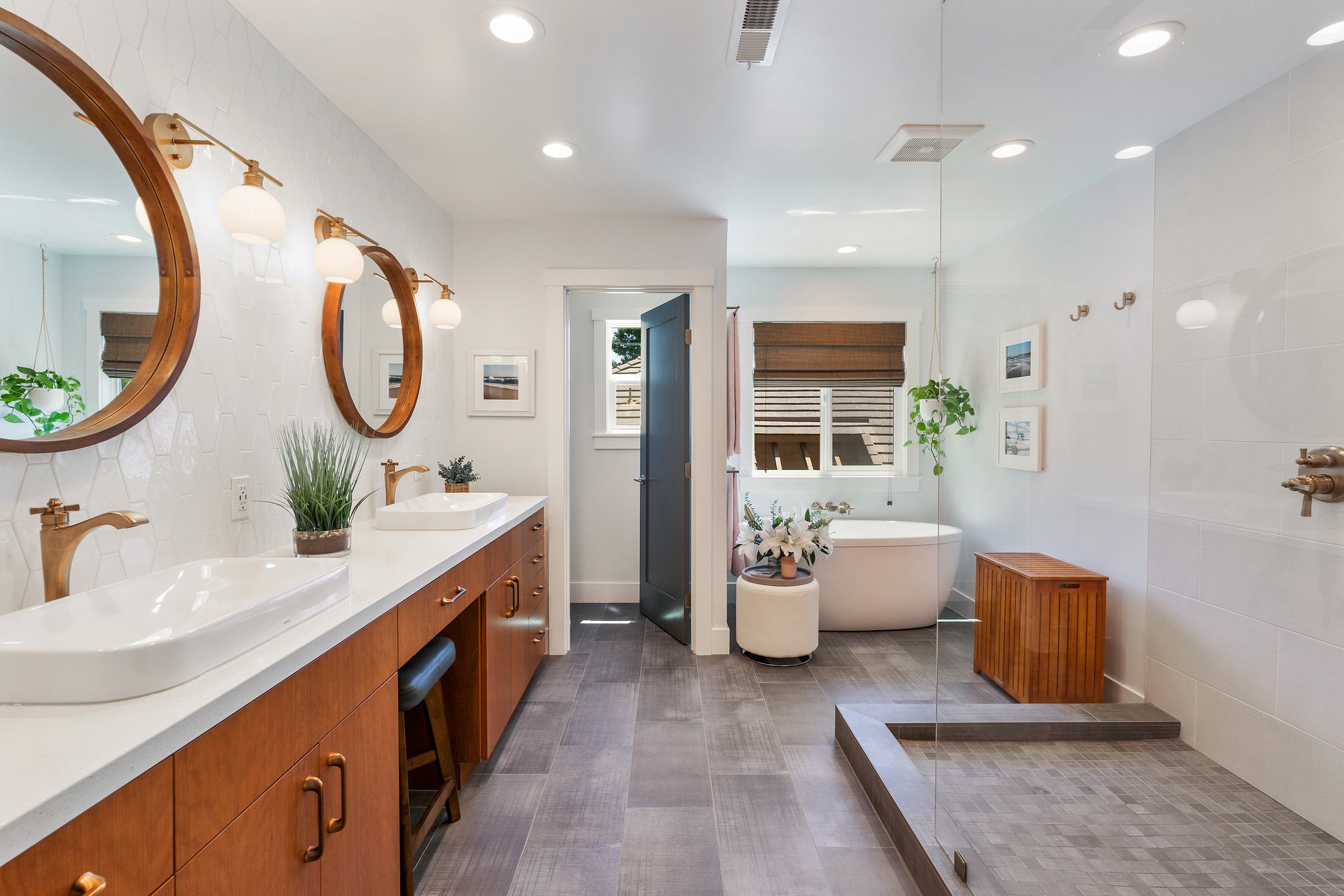 Modern bathroom with double vanity sinks, round mirrors, brass fixtures, potted plants, a bathtub, a wooden storage bench, and a walk-in shower with gray tiled flooring and white walls.
