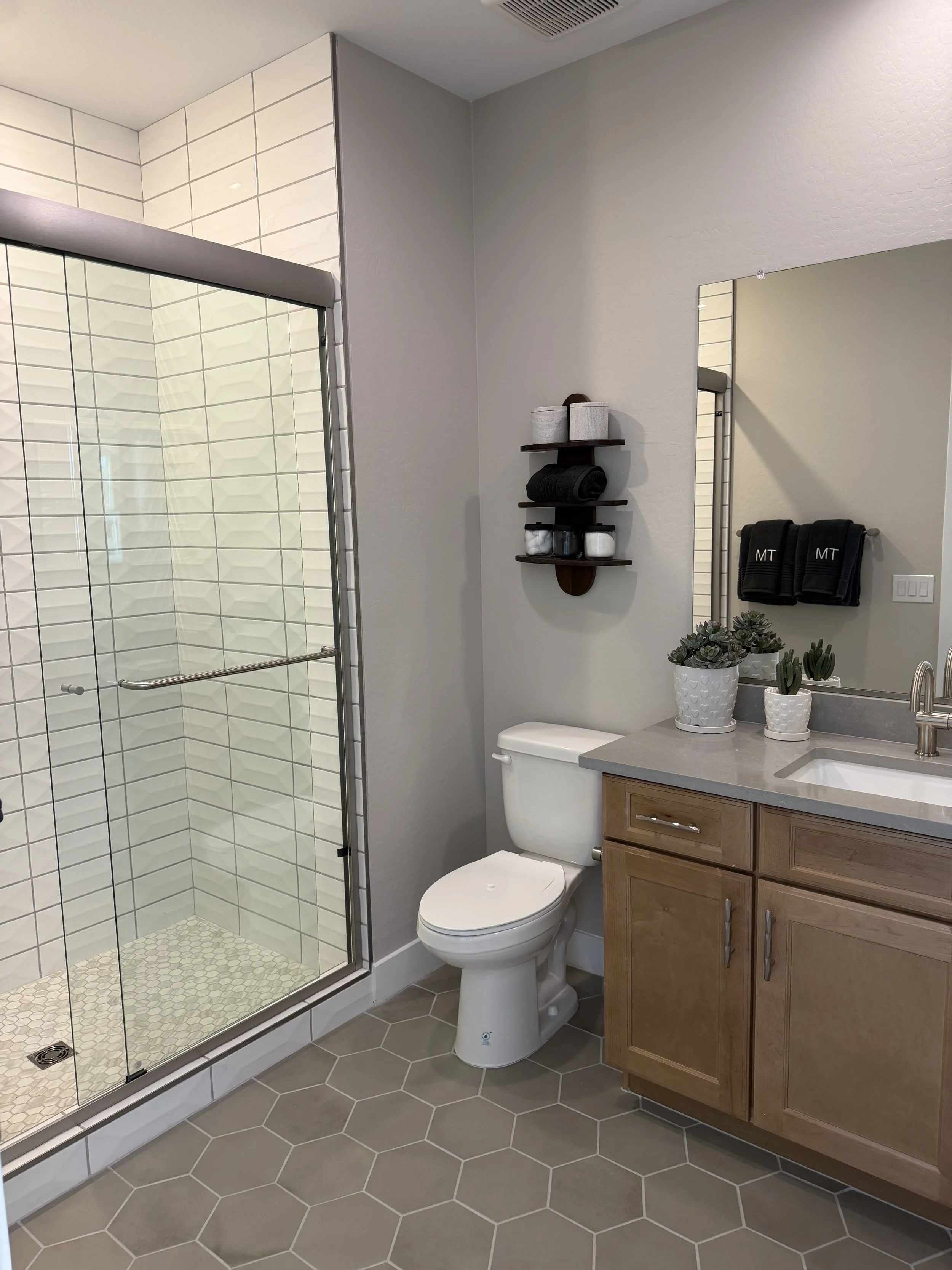 Modern bathroom with glass shower, toilet, wooden vanity with gray countertop, potted plants, and wall-mounted towel rack.