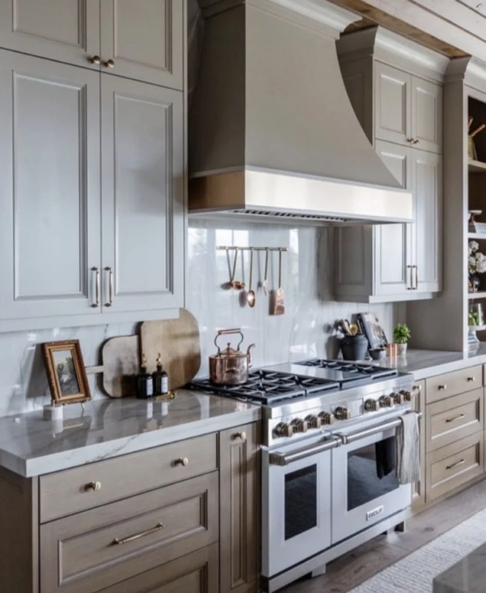 Kitchen with gray cabinetry, marble countertop, stainless steel stove, oven, and range hood, decorated with picture frames, cutting boards, plants, and copper utensils.
