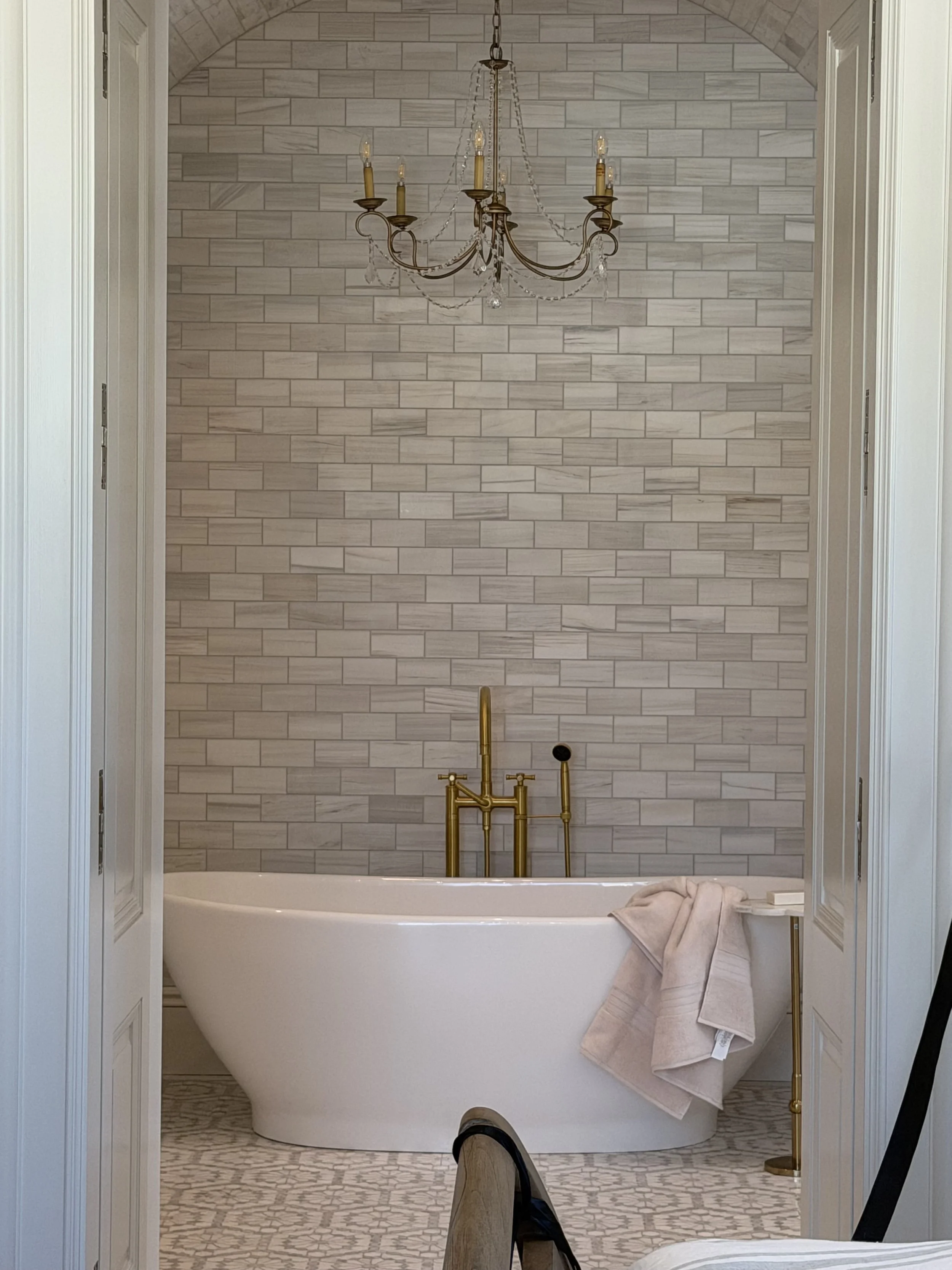 A bathtub with gold fixtures in front of a beige brick tile wall, with a towel draped over the edge of the tub and bathroom furniture visible around it.