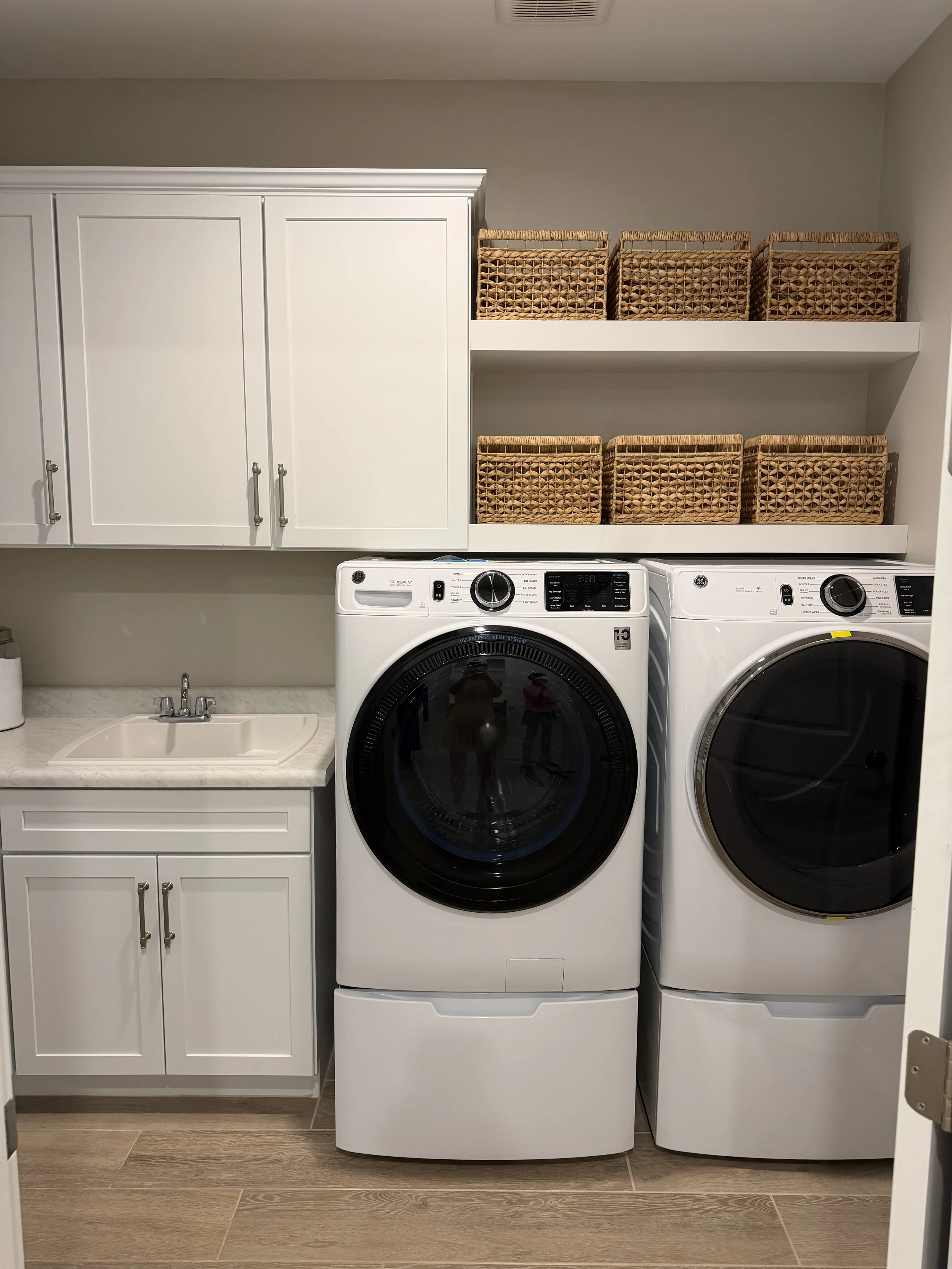 A laundry room with white cabinets, a small sink, a washer, and a dryer. Wicker baskets are stored on a wall shelf.
