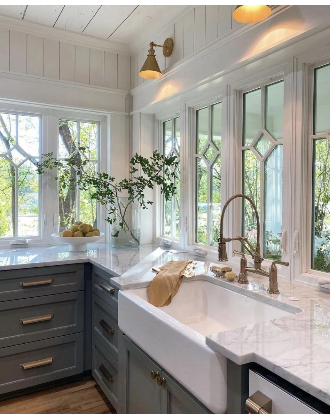 Bright kitchen with three large windows, white walls, and a marble countertop. A vase with green branches and a bowl of apples sit on the counter. Gold fixtures are on the faucet and lighting, and there is a beige towel near the sink.
