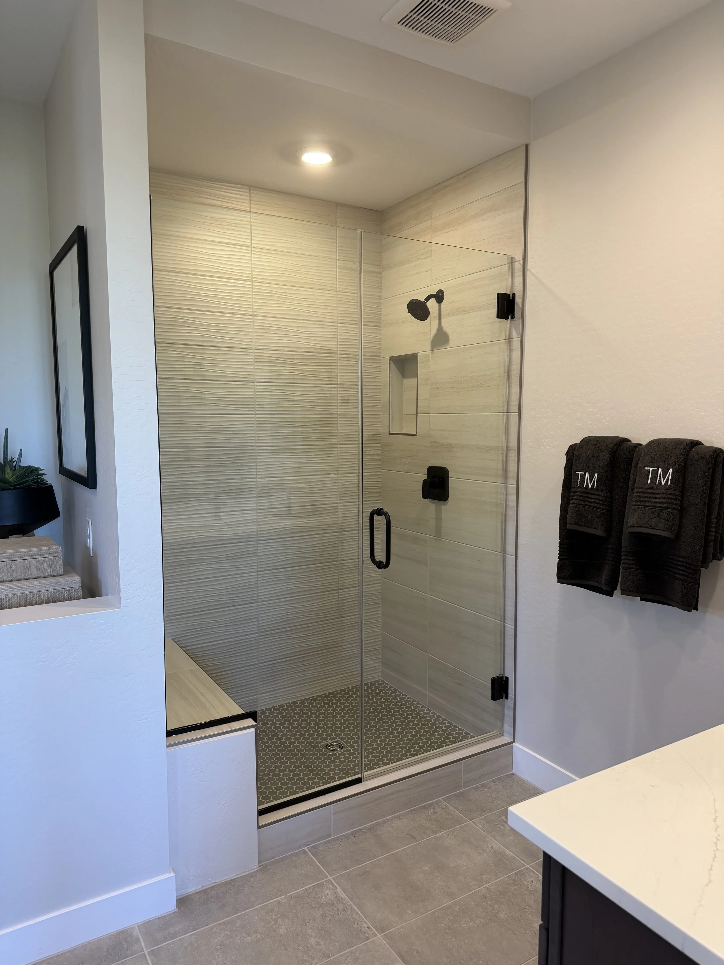 A modern bathroom with a glass-enclosed walk-in shower featuring beige textured and smooth tiles, a black showerhead, and a small built-in shelf. Black towels with white monogrammed initials hang on a rack on the right wall. The bathroom has gray flo