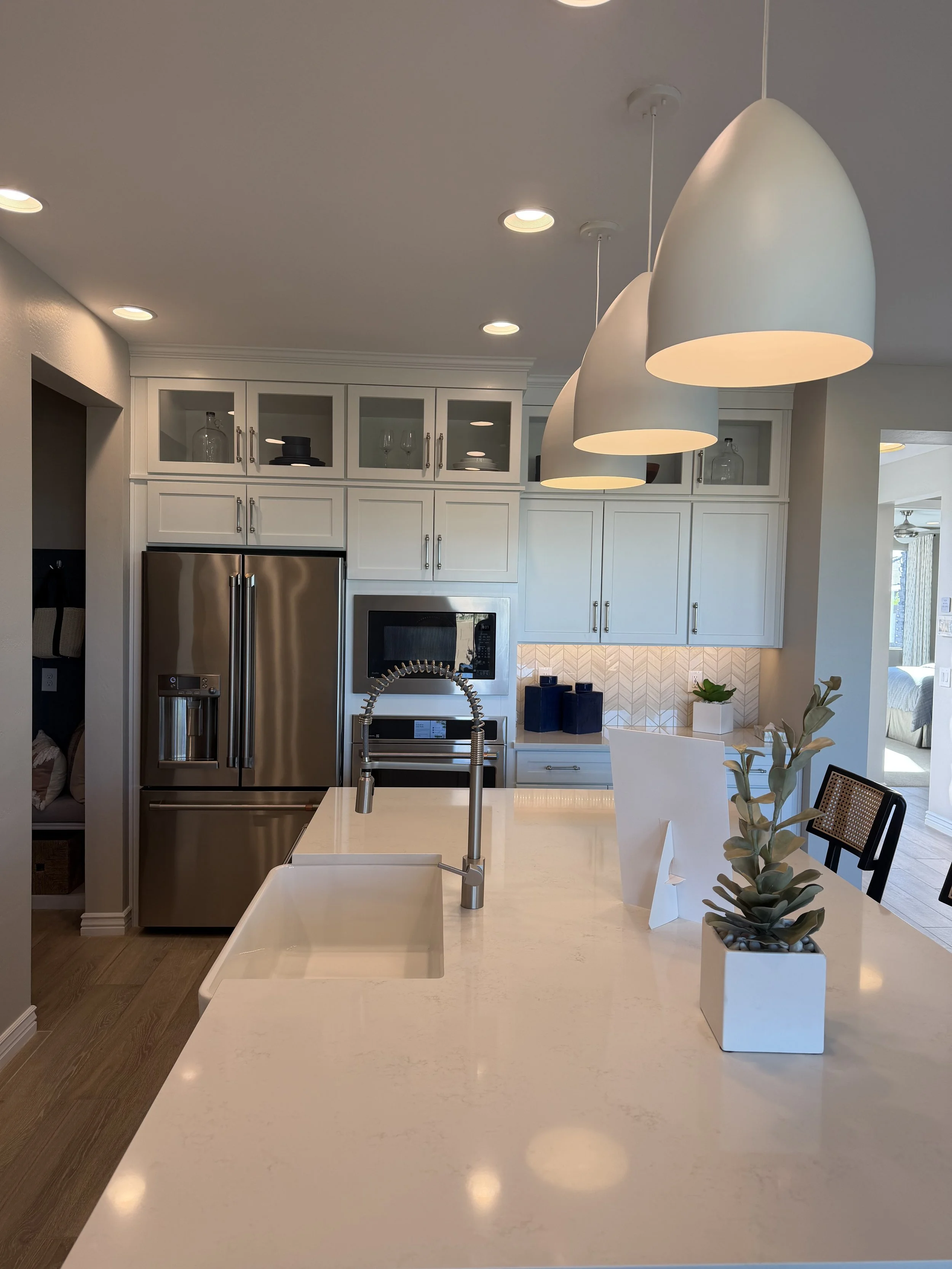 Modern kitchen with a white island countertop, a white cabinet with glass doors, a stainless steel refrigerator, a microwave oven, pendant lights, and a flower arrangement in a white vase on the counter.