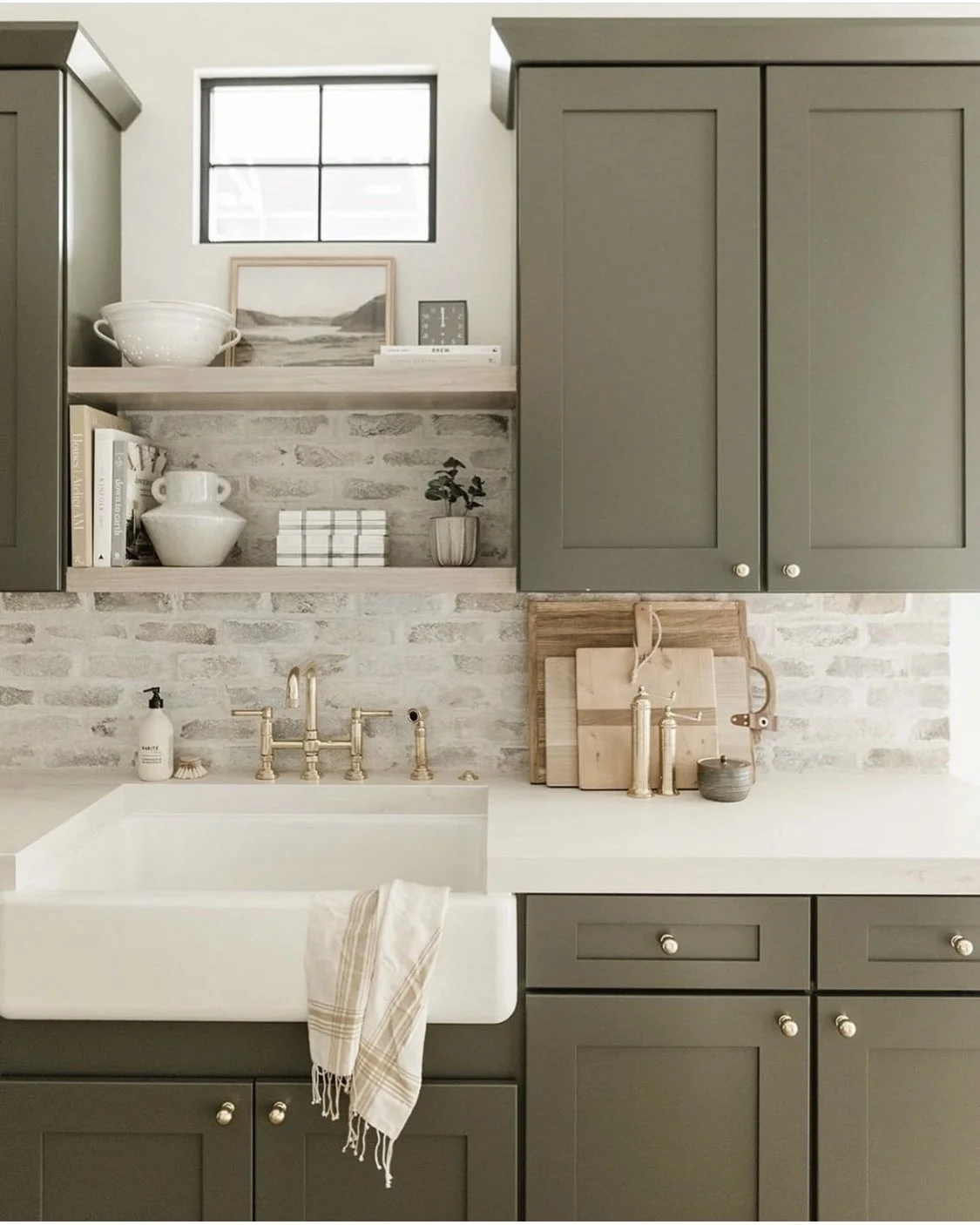 Kitchen with green cabinets, a white farmhouse sink with a beige towel, open shelves with dishes and decor, a small window, and a white brick backsplash.