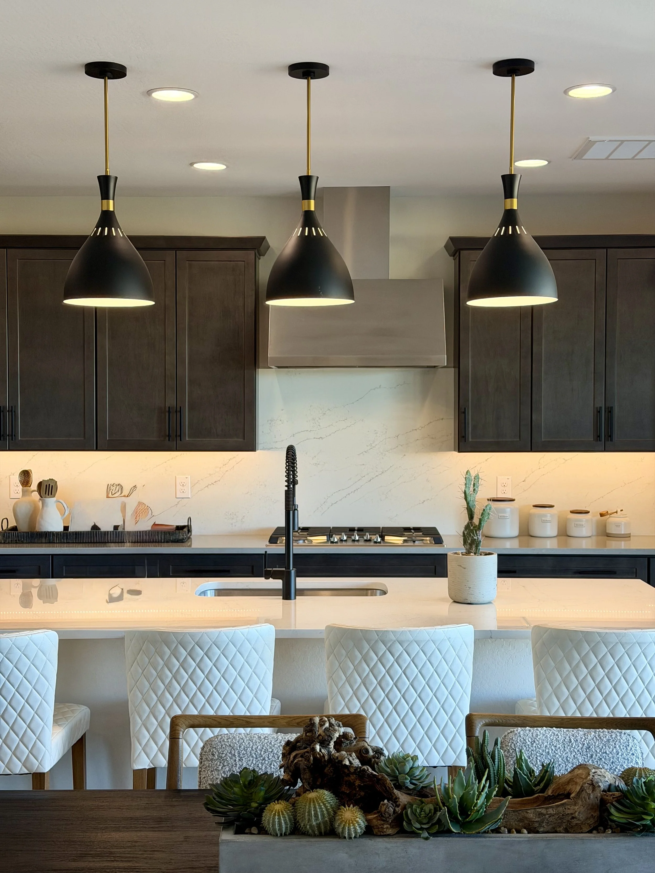 Modern kitchen with hanging black pendant lights, dark wooden cabinets, white marble backsplash, island with sink, potted cactus, and white quilted chairs.