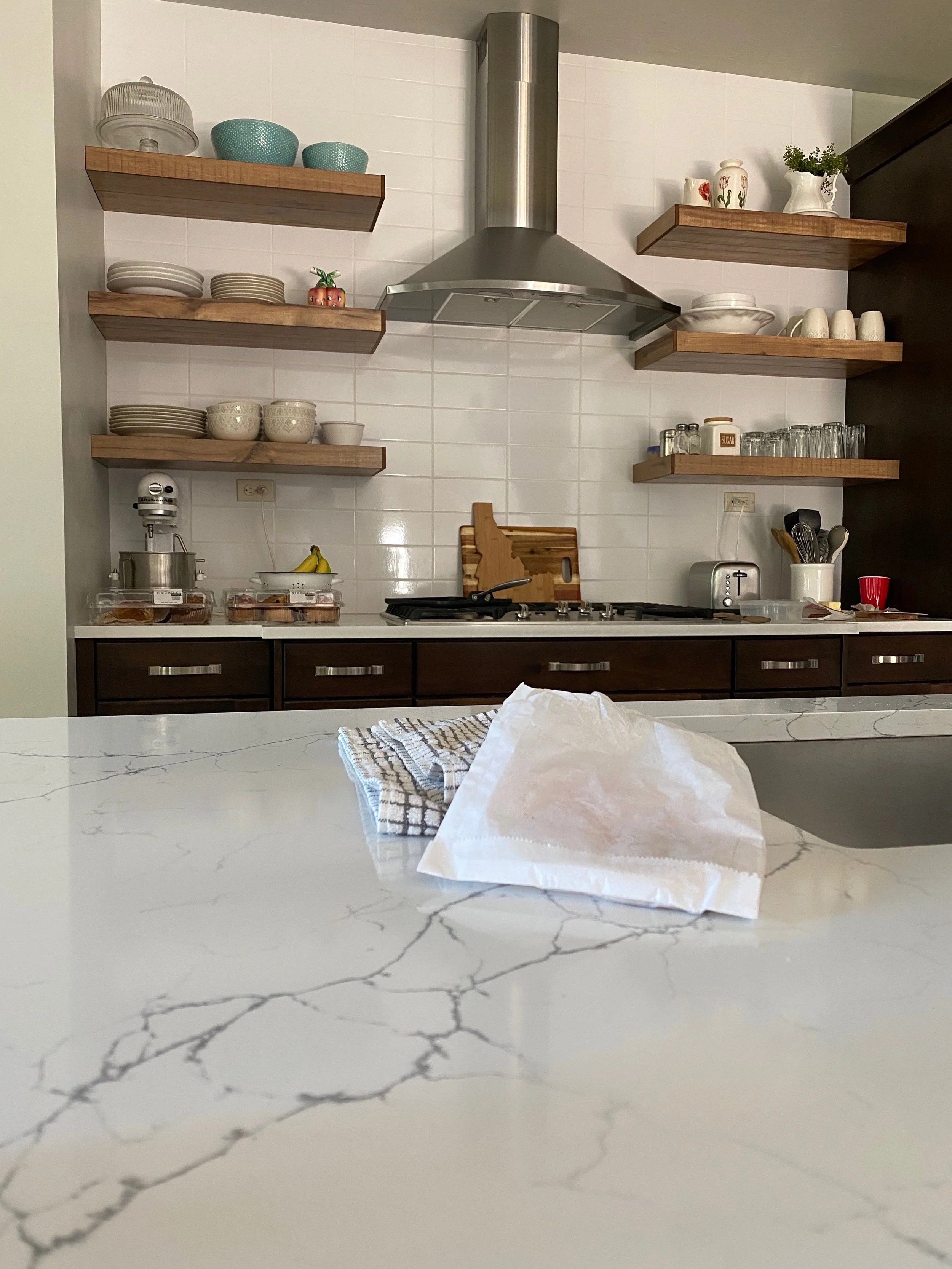 Kitchen countertop with a paper bag and towel, with shelves of dishes, bowls, glasses, and kitchen utensils in the background.