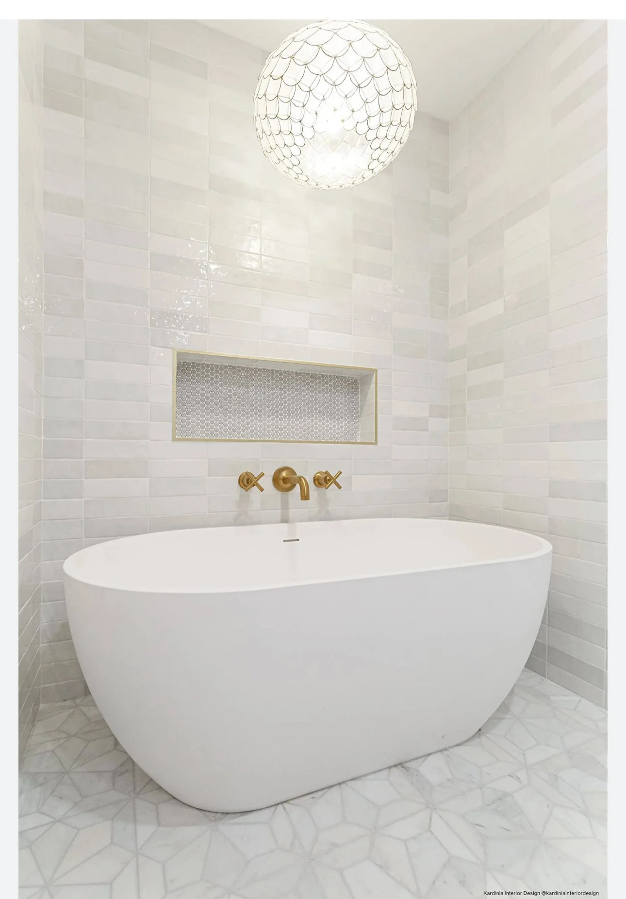 A modern white freestanding bathtub in a minimalist bathroom with light gray tiled walls, a built-in shelf, and a ceiling light fixture.