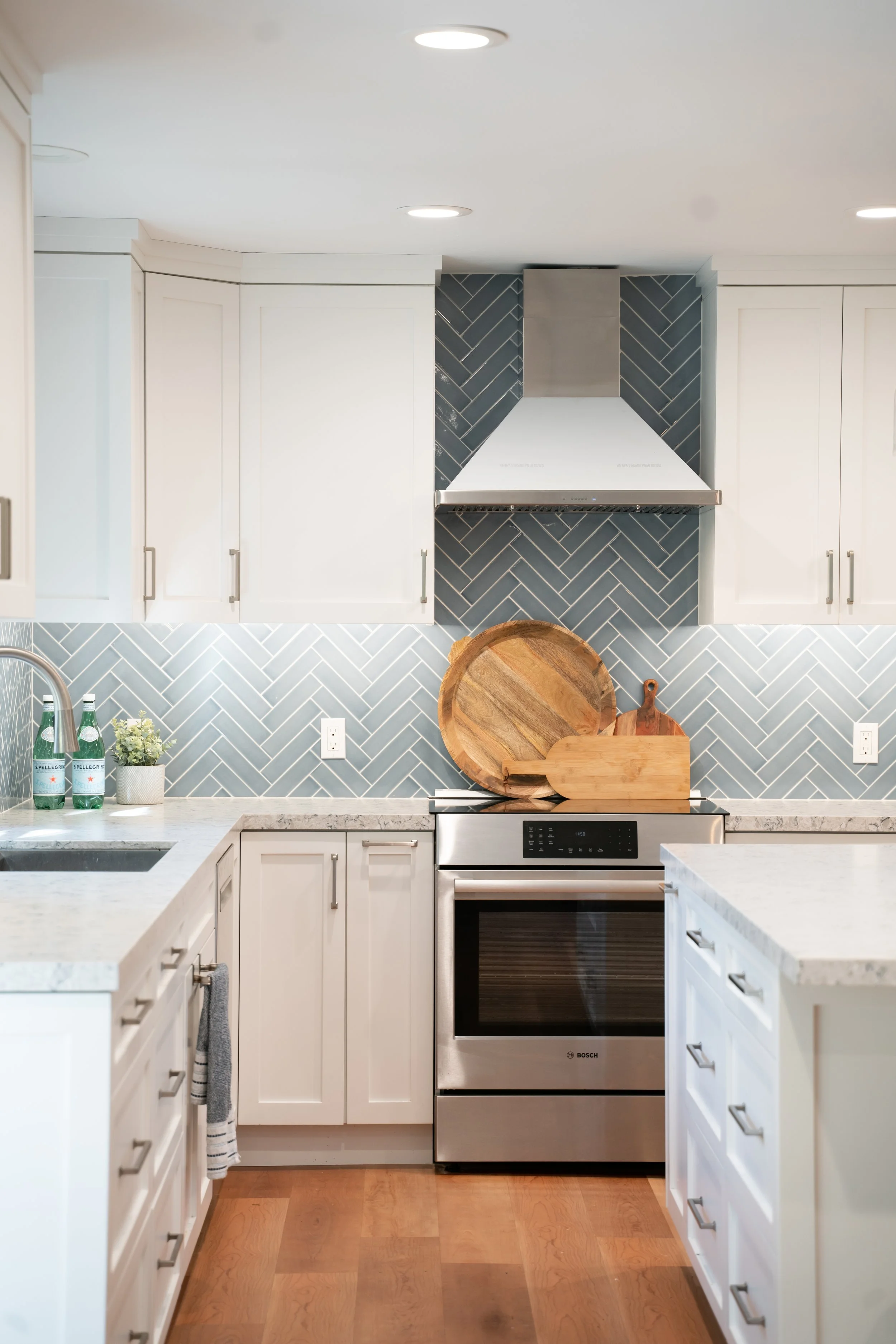 Modern kitchen with white cabinets, stainless steel oven, gray herringbone tile backsplash, wooden cutting boards, and two bottles of sparkling water on the counter.