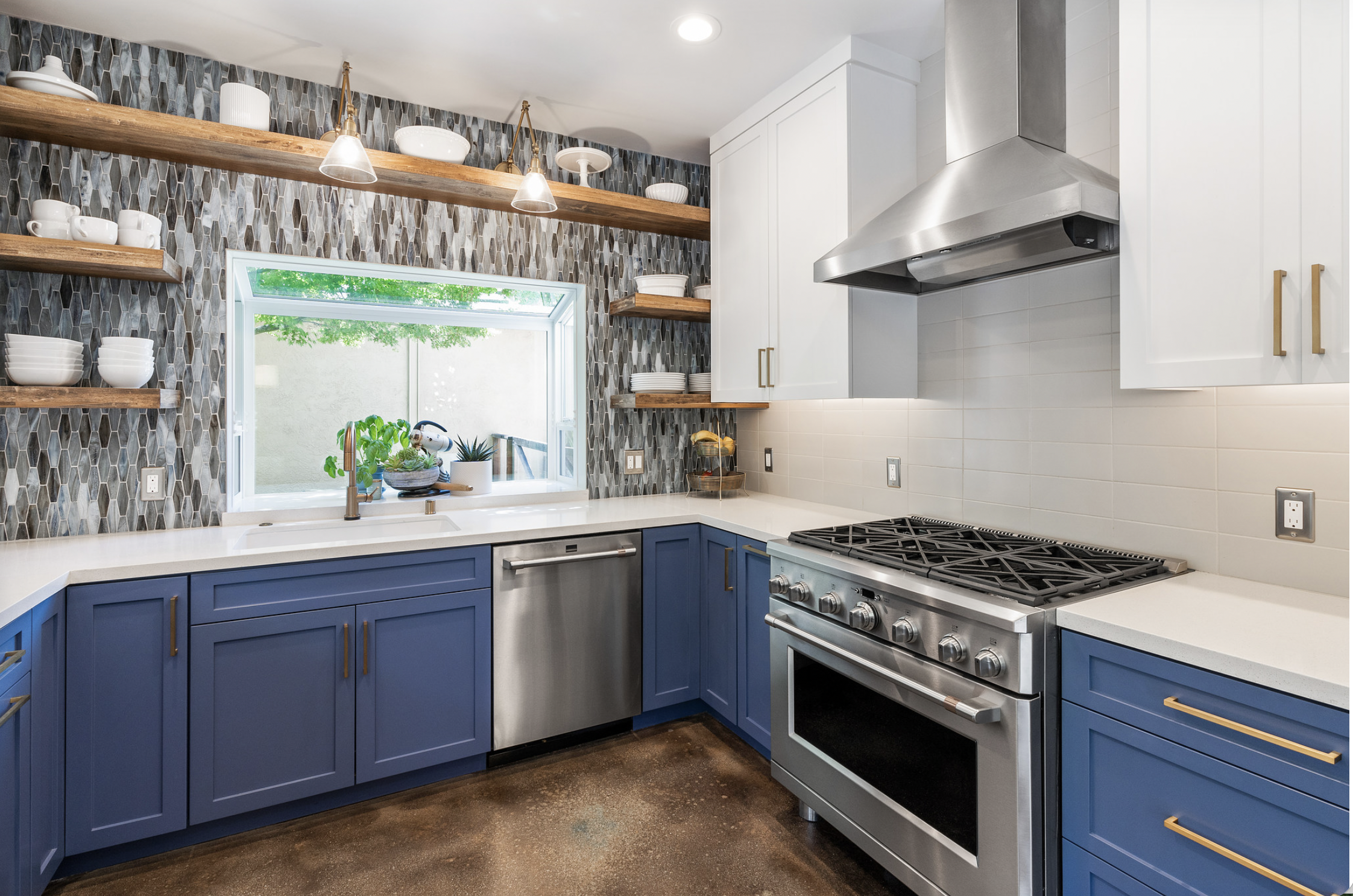 Modern kitchen with blue lower cabinets, white upper cabinets, white countertops, stainless steel appliances, open wooden shelves, a window with greenery outside, decorative backsplash tiles, and a polished concrete floor.