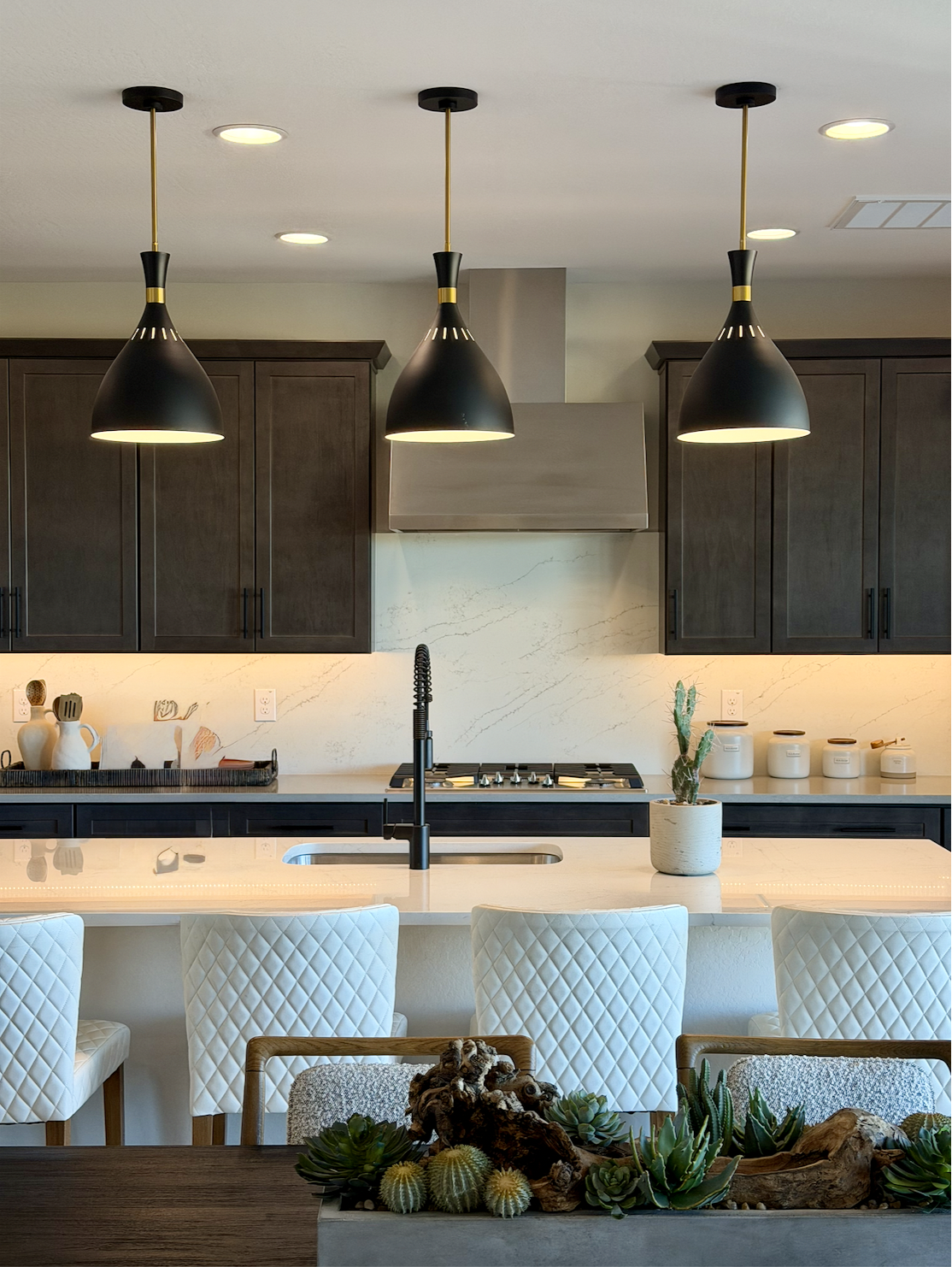 Modern kitchen with dark cabinets, marble backsplash, and three black hanging pendant lights over a white island with a black faucet, white quilted chairs, and decorative plants.