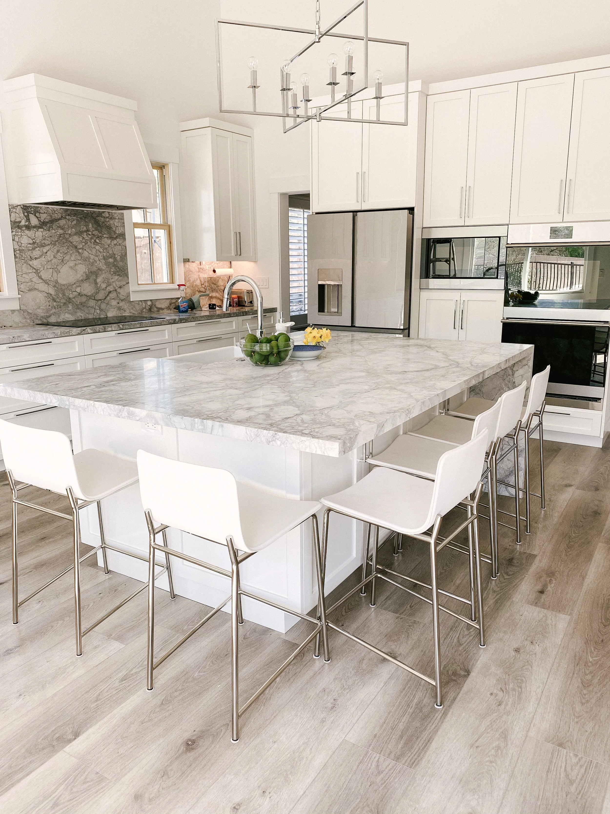 A modern kitchen with white cabinets, a marble island, and bar stools, featuring stainless steel appliances and a light wooden floor.