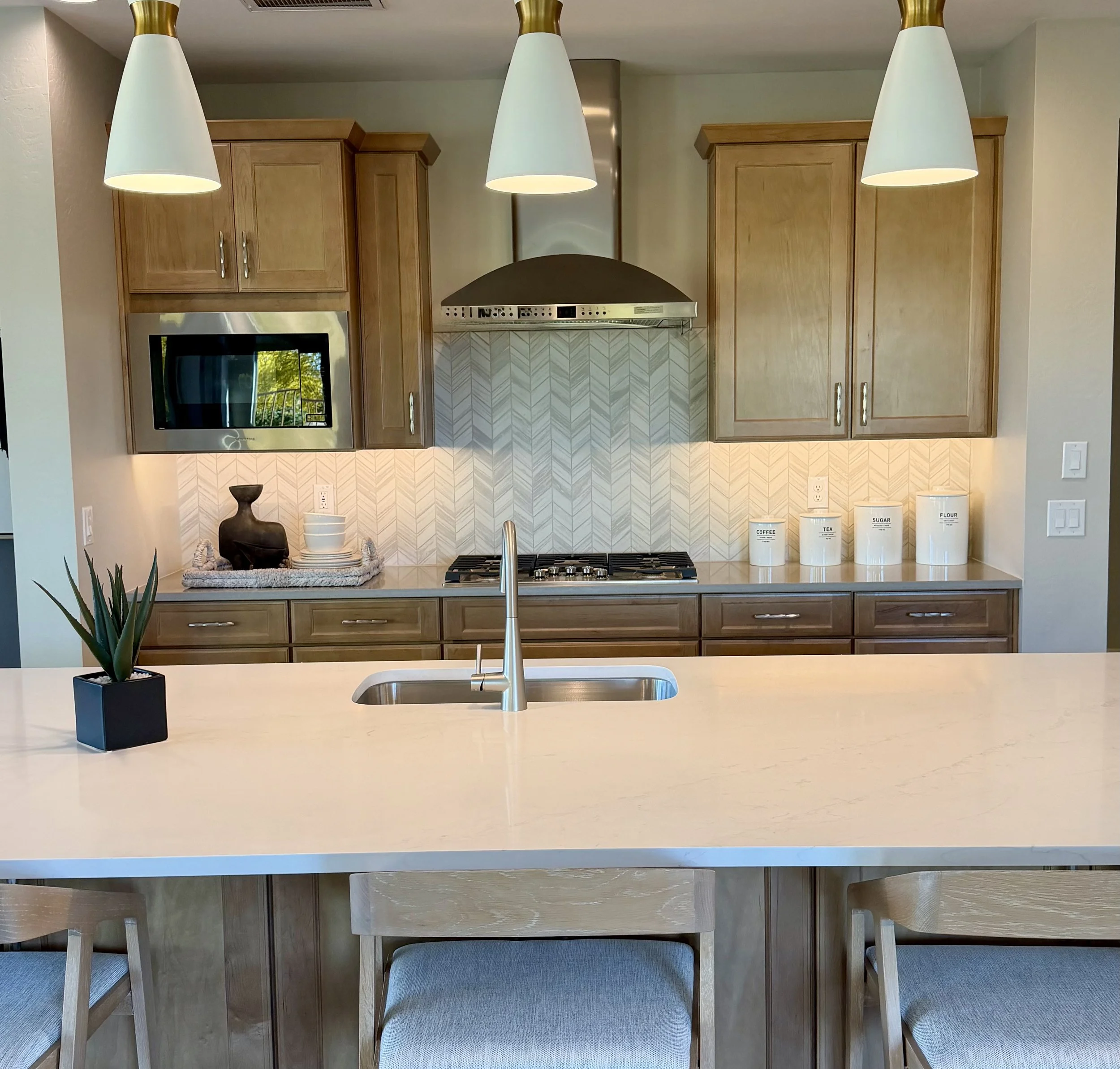 Modern kitchen with light wood cabinets, a white marble island, pendant lights, stainless steel range hood, microwave, and canisters for coffee, tea, sugar, and flour.