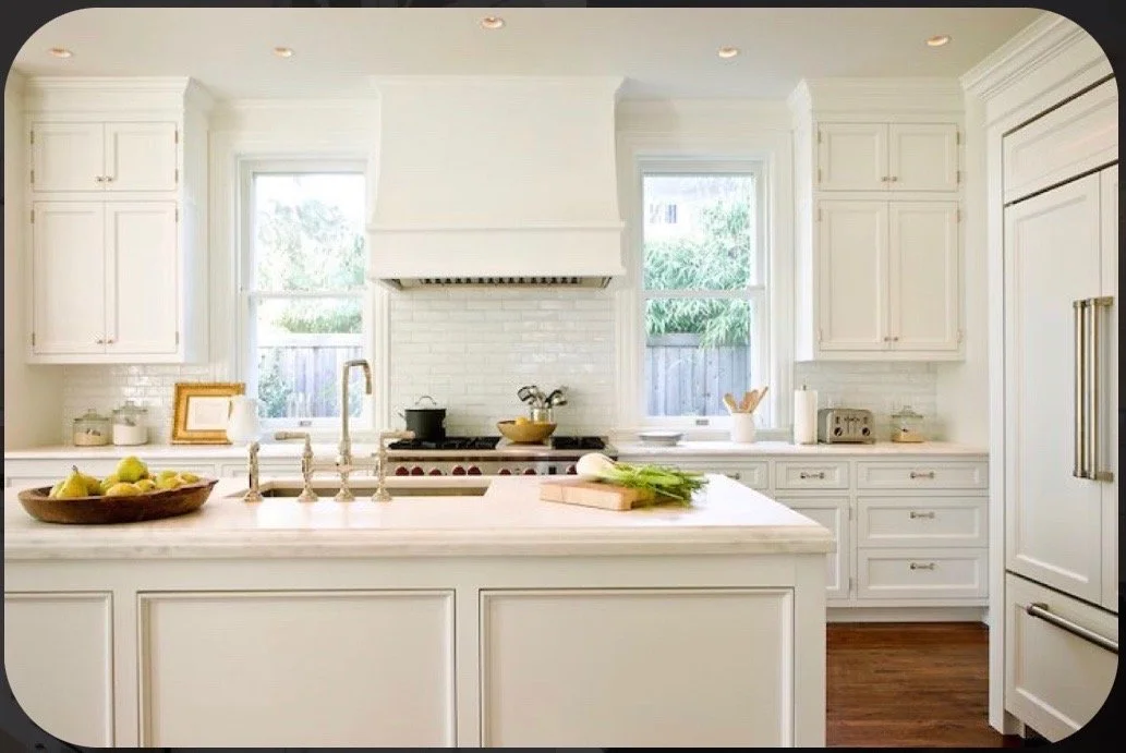 Bright, white kitchen with center island, stainless steel appliances, and natural light from two windows, featuring fresh produce and cooking utensils.