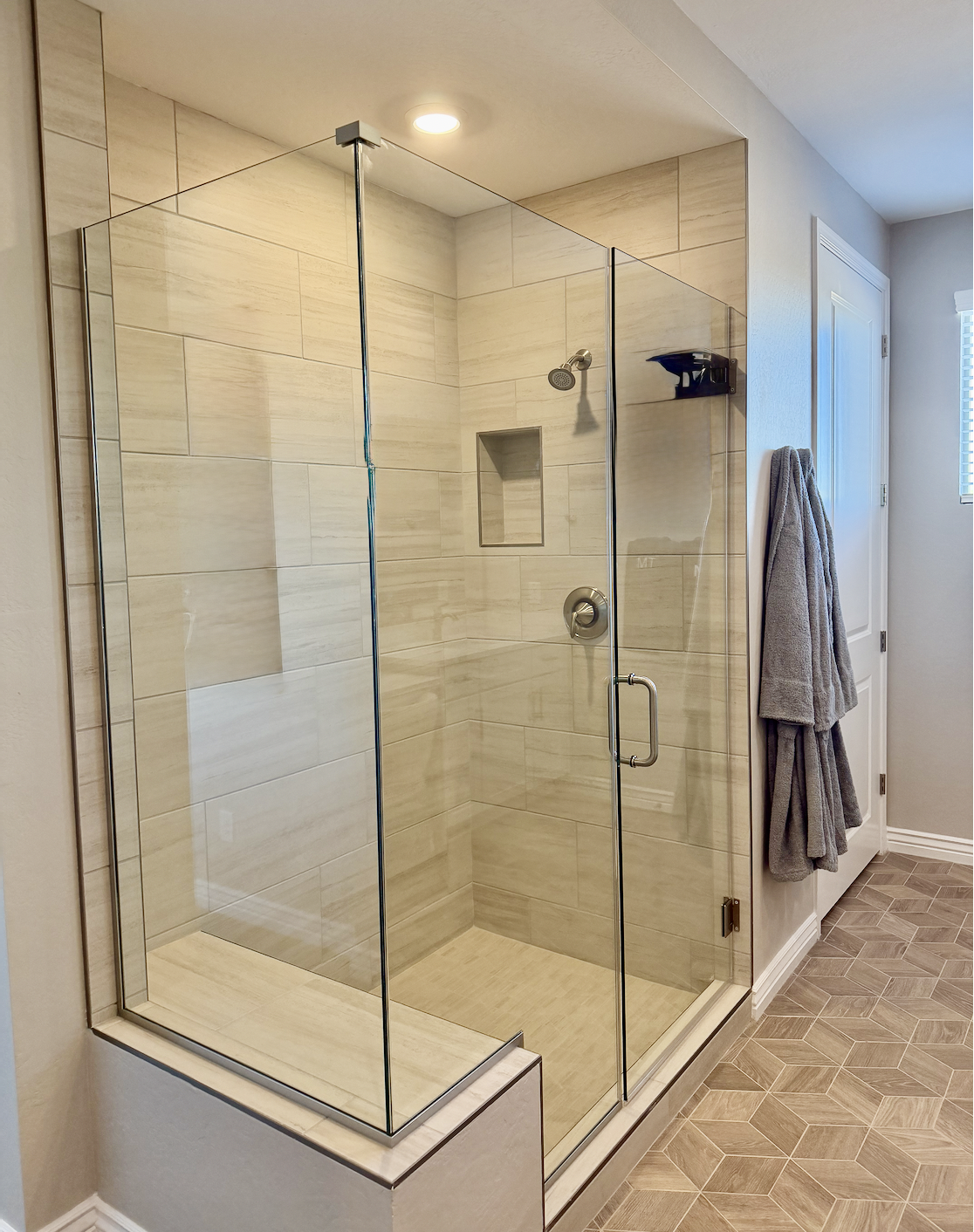 Empty walk-in shower with glass doors, beige tiles, built-in niche, and a gray towel hanging on a hook nearby.