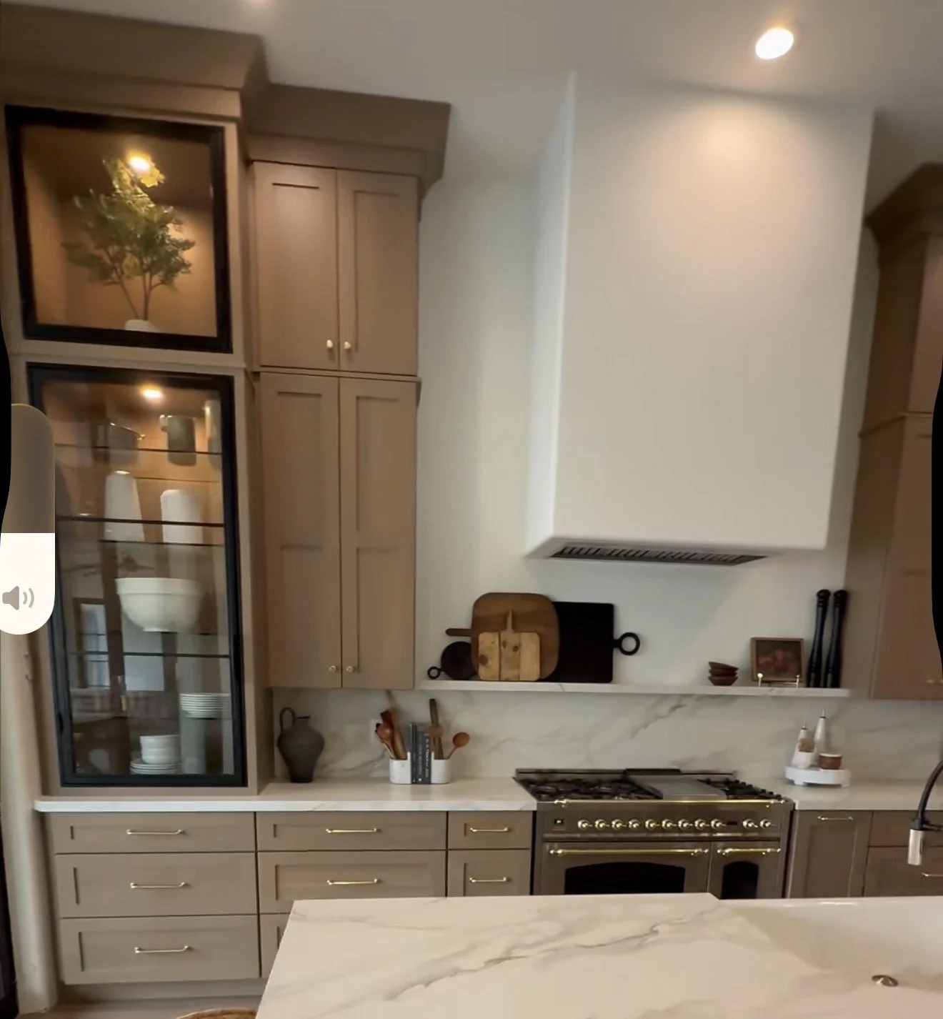 Modern kitchen with beige cabinets, white marble countertops, a stainless steel stove, and a wall-mounted white range hood. Decorative items include a cutting board, pitchers, and small bowls on the shelf.