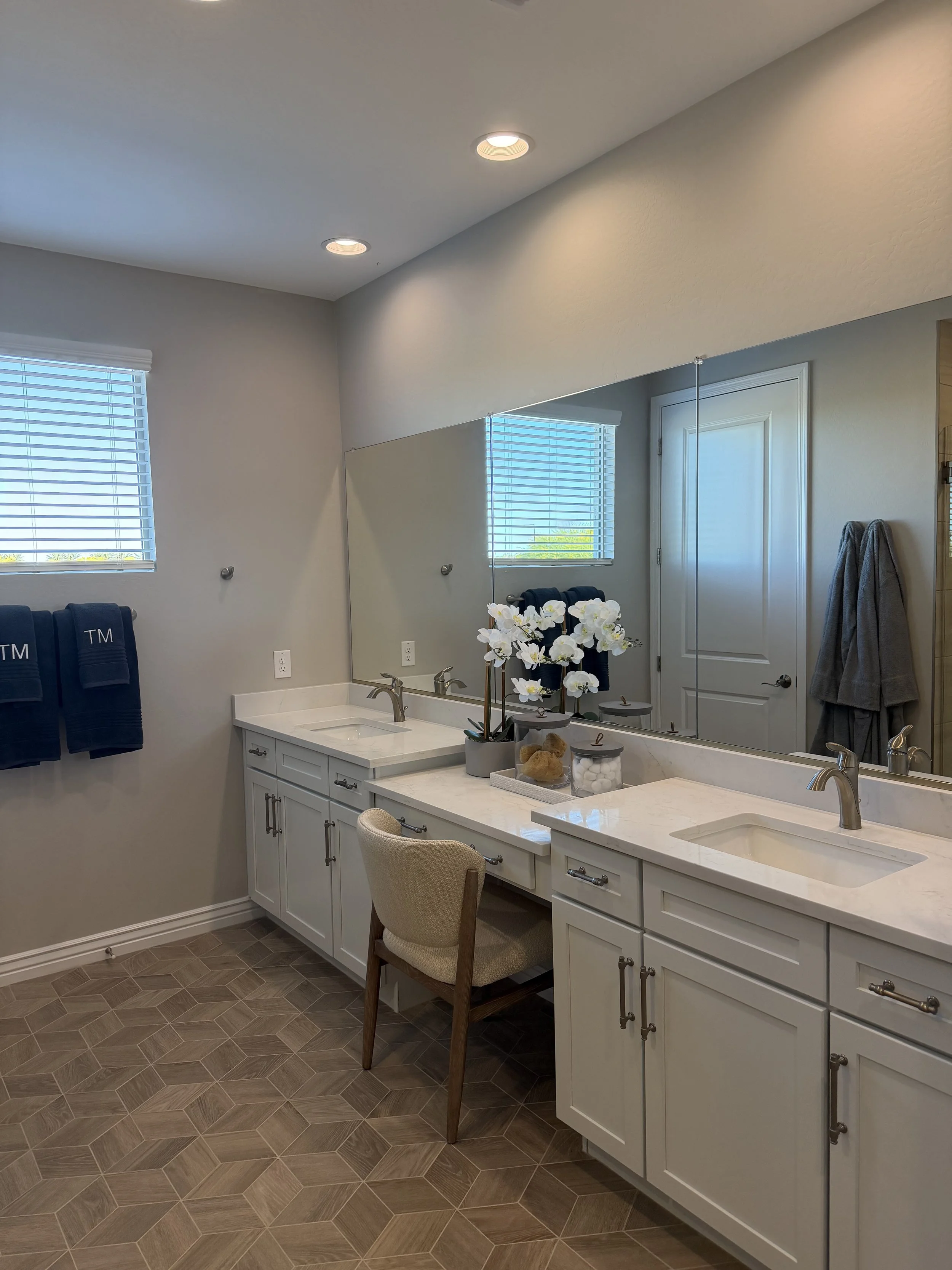 A bathroom vanity with a white countertop and cabinets, a sink, a chair, a mirror, and a decorative flower arrangement, with towel holders and windows in the background.