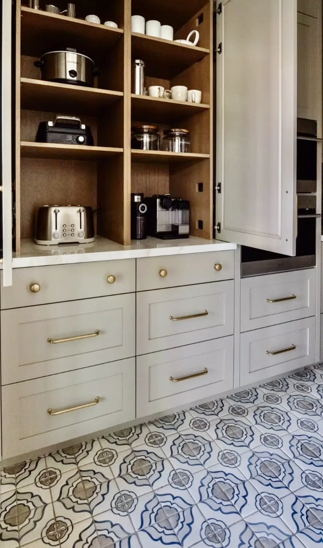 Kitchen cupboard with shelves containing appliances such as a toaster, coffee maker, and rice cooker; the lower cabinets have gold handles and the kitchen floor has patterned tiles.