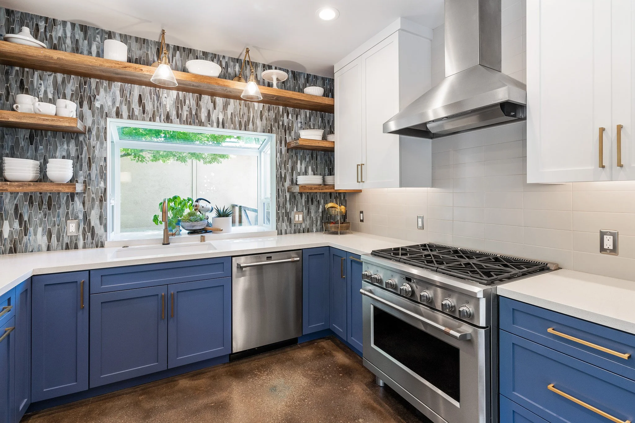Modern kitchen with blue cabinets, stainless steel stove, white countertops, open wooden shelves with dishes, decorative tiled backsplash, window with plants outside, and a bright, clean design.