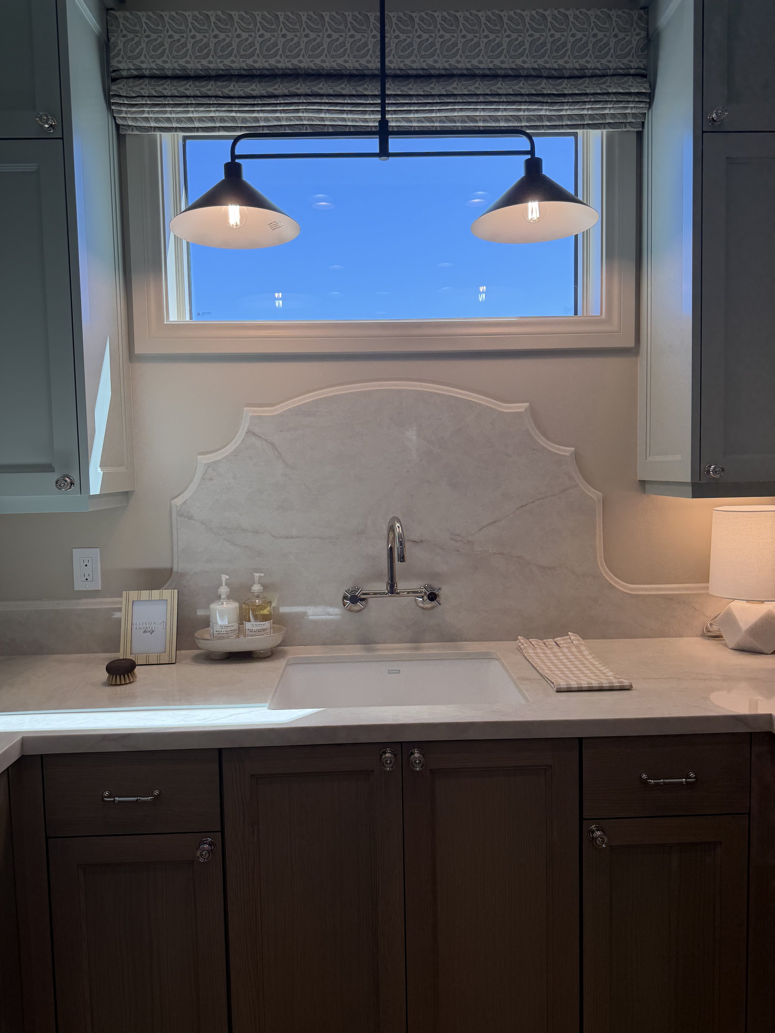 Kitchen sink area with a window above it, showing a blue sky outside. The countertop has a soap dispenser, a small framed picture, a brush, and a folded cloth. There is a lamp with two shades hanging from the ceiling.