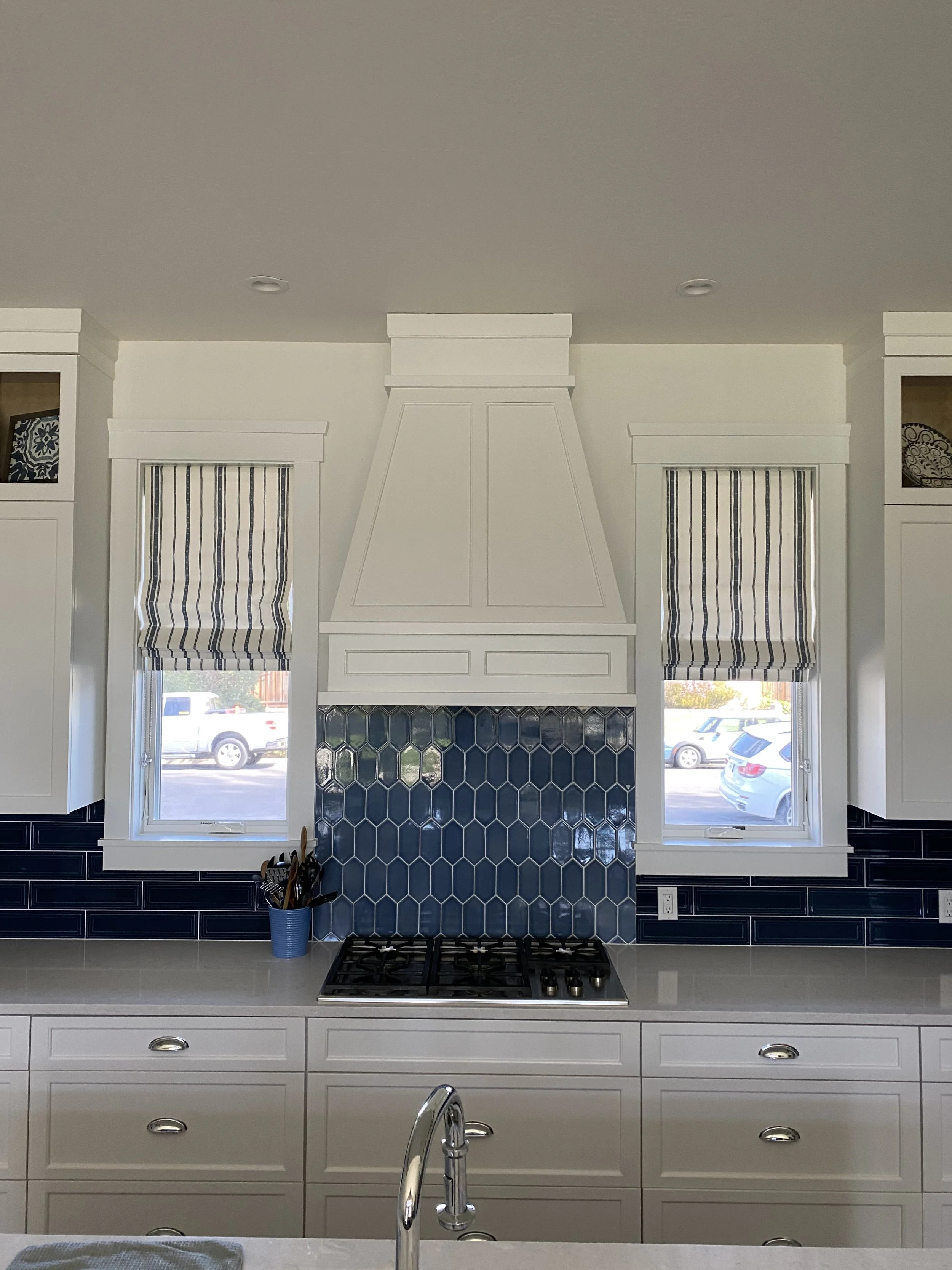 A modern kitchen featuring white cabinetry, a stovetop, blue hexagonal tile backsplash, and two windows with striped valances.