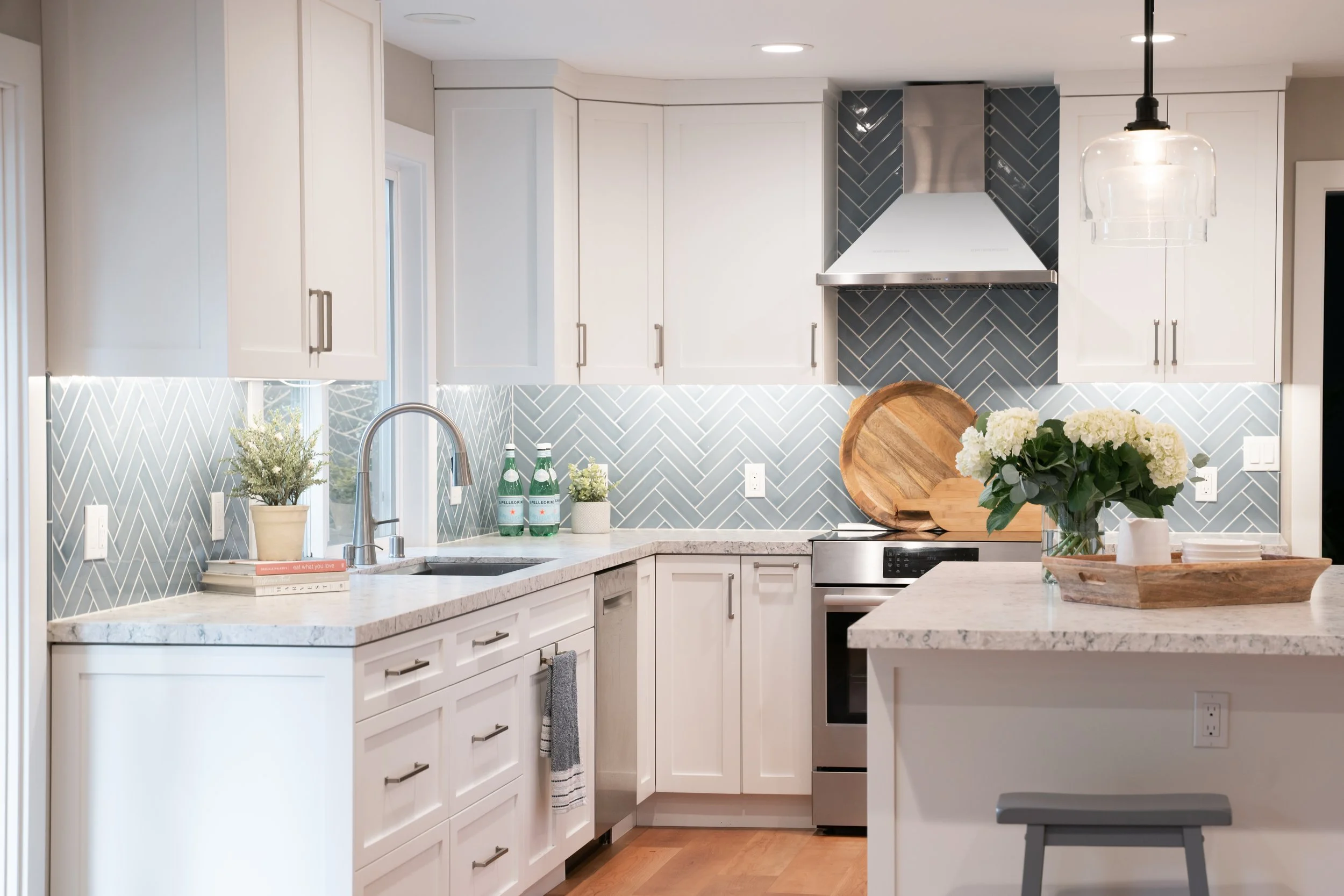 Modern kitchen with white cabinets, pastel blue backsplash in a herringbone pattern, granite countertops, stainless steel appliances, and decor including potted plants, bottled sparkling water, and a vase of white flowers.