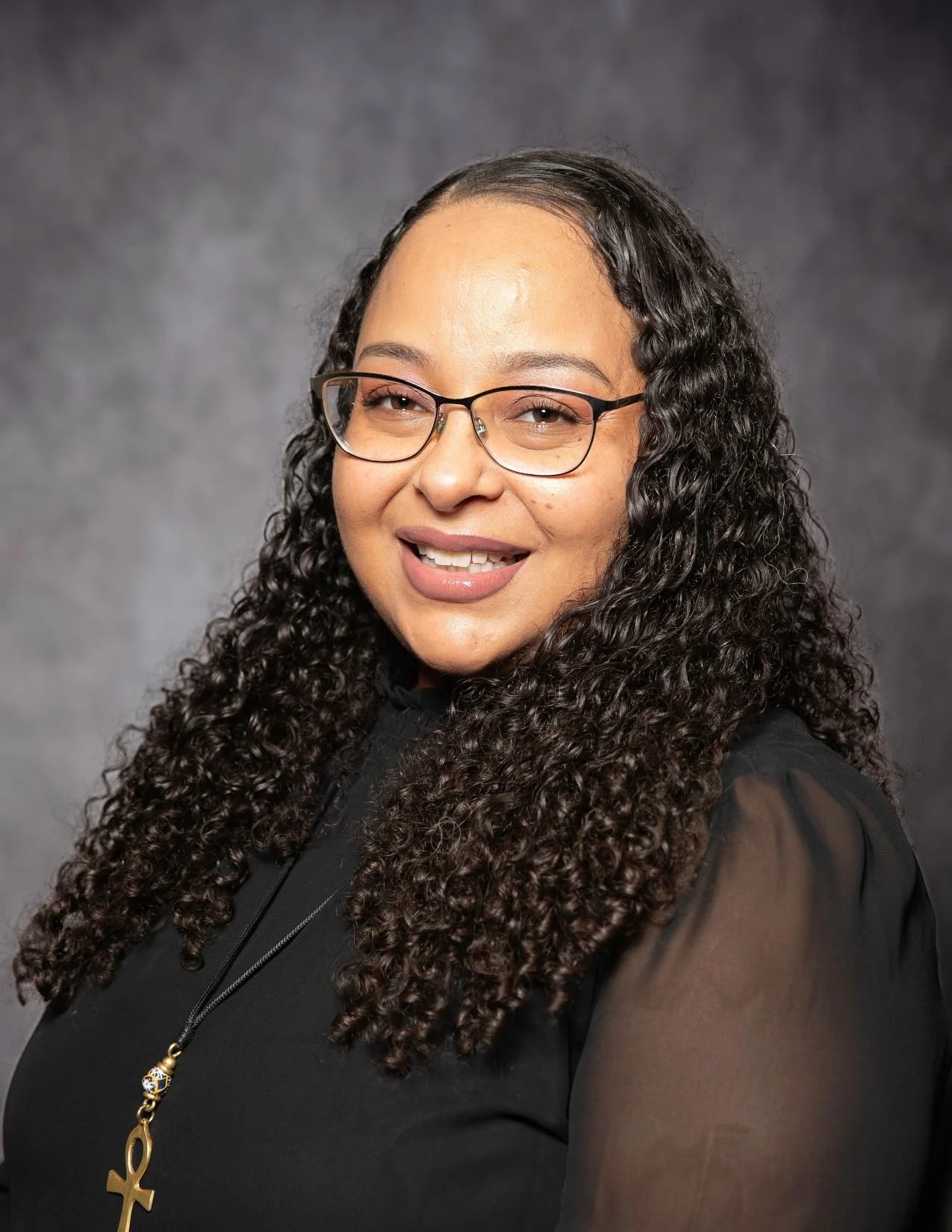 portrait of a woman with curly dark hair, glasses, and a black top, smiling against a gray background.