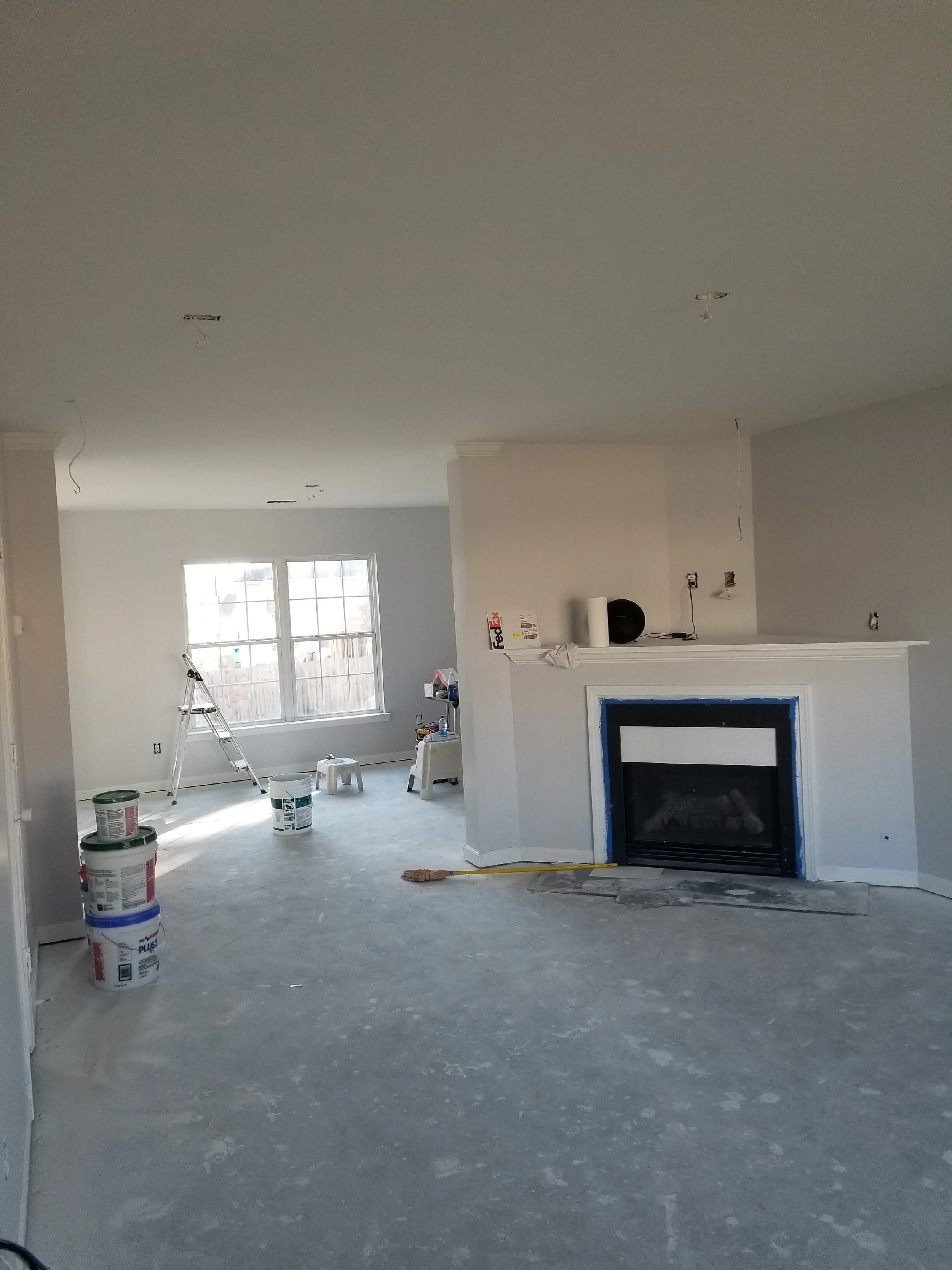 Living room under renovation with a fireplace, partially painted walls, and construction supplies including cans of paint, ladder, and tools.