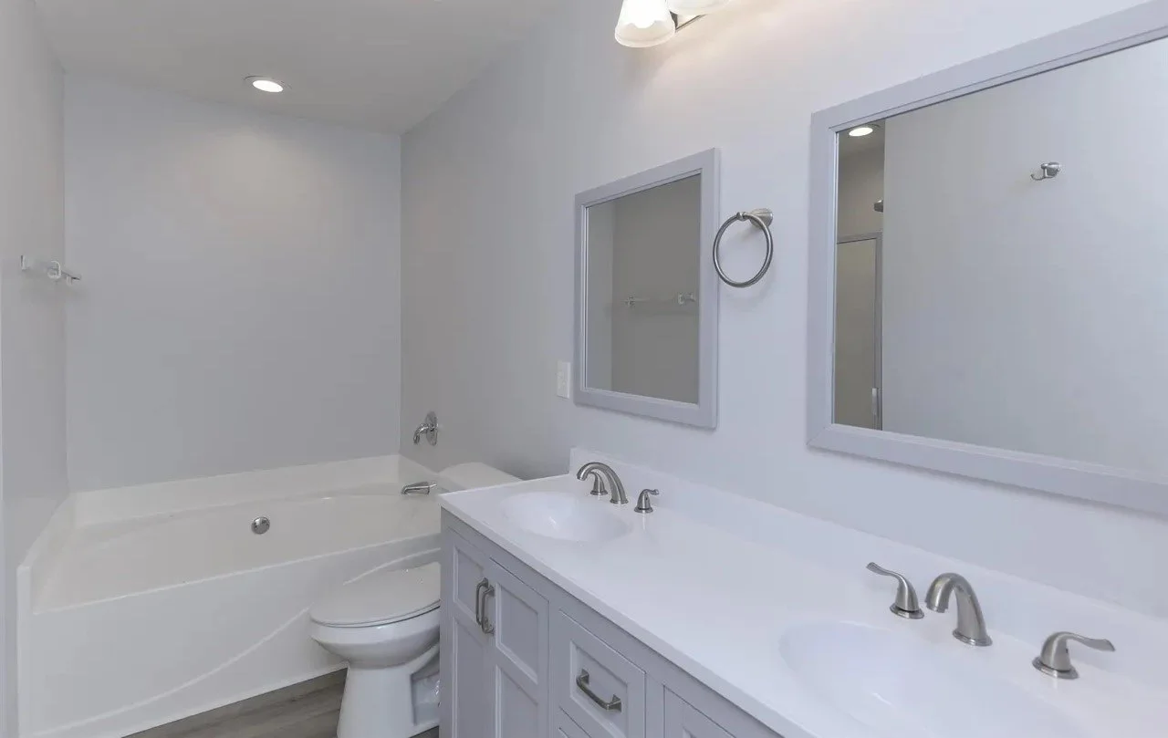 A cleaned and renovated modern bathroom with a white vanity featuring two sinks, two mirrors, a bathtub, a toilet, and gray accents.