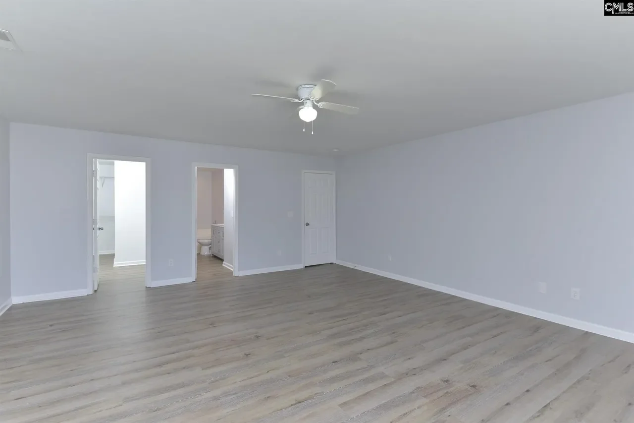 Renovated now empty room with light gray walls, light wood flooring, a ceiling fan with lights, and two doorways leading to a bathroom and a closet.