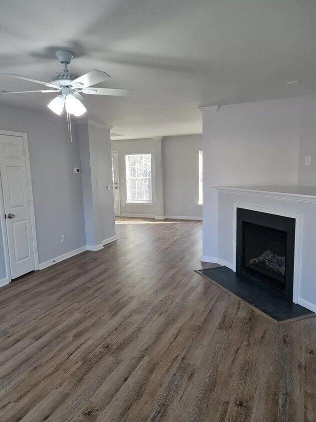 Empty living room with wood flooring, white walls, a white ceiling fan, and a fireplace with a black surround.