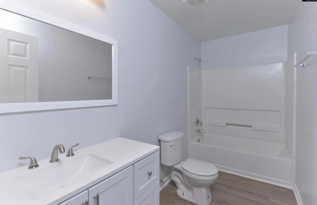 A cleaned and renovated white bathroom with a mirror, vanity with faucet, toilet, and bathtub with showerhead.