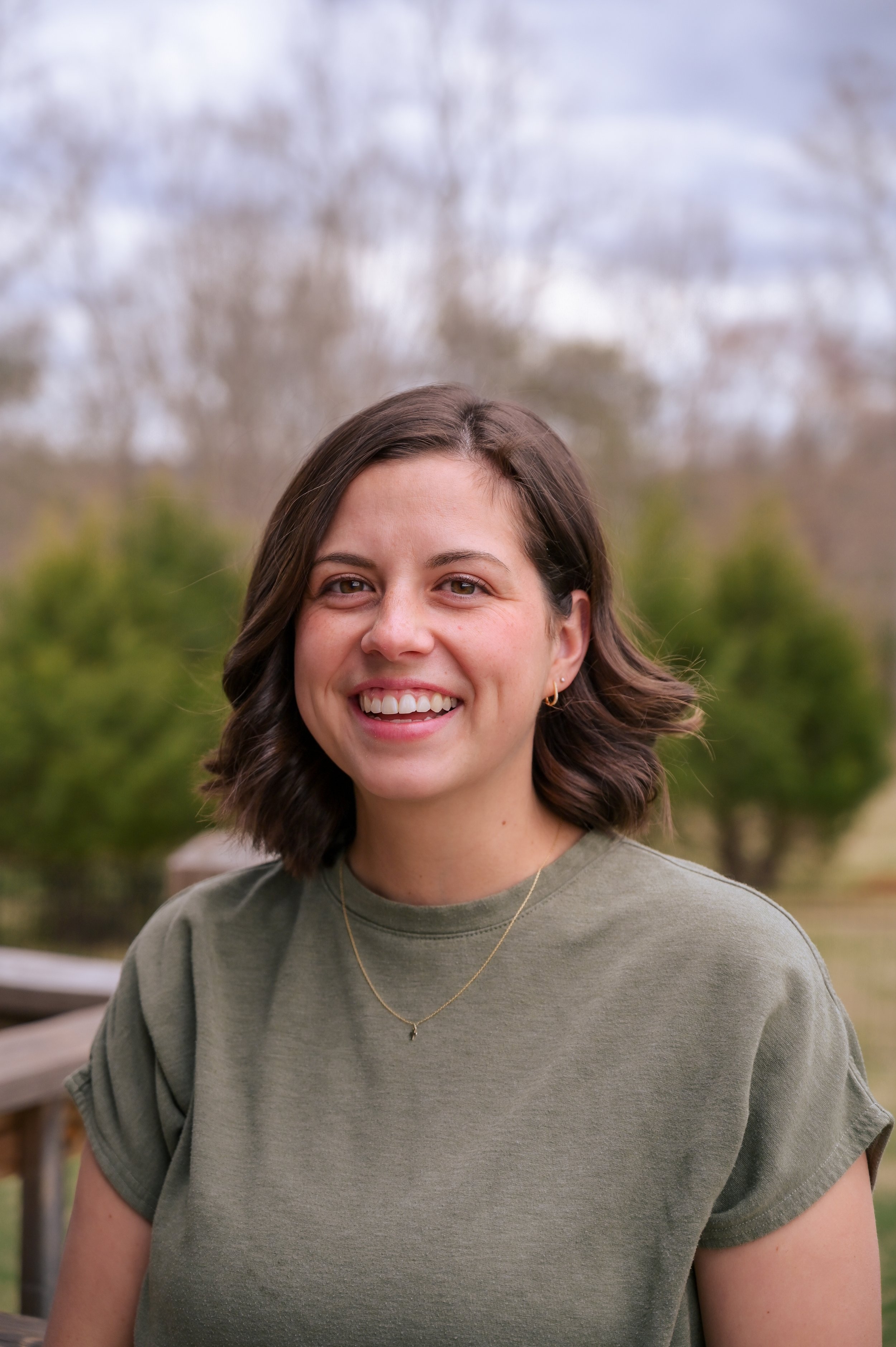 A young woman with wavy brown hair, smiling, wearing a green t-shirt and a gold necklace, standing outdoors on a cloudy day with trees in the background.