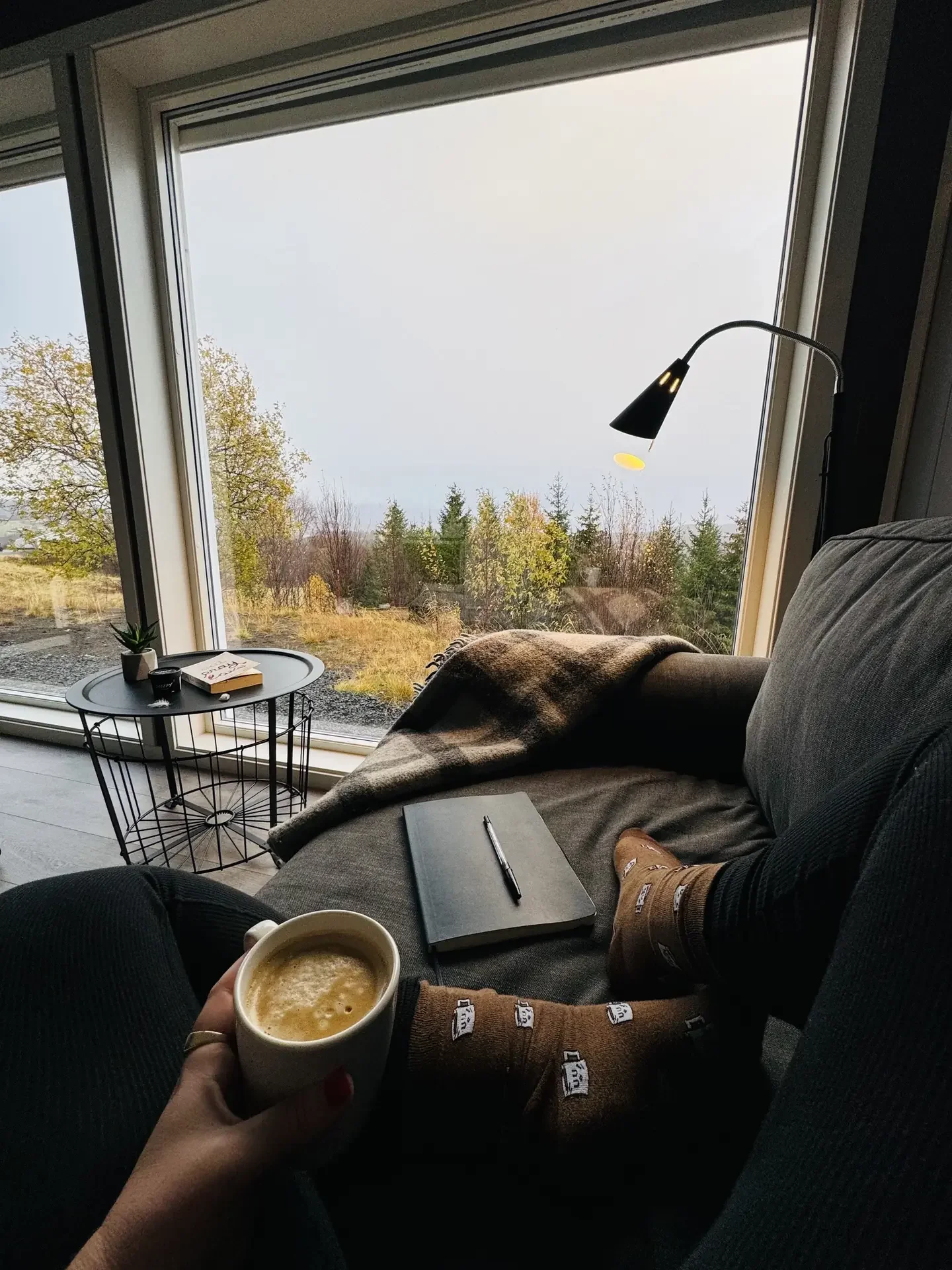 Relaxing in a cosy armchair with a coffee and an open journal, wrapped in a blanket beside a large window looking out over an autumn forest.