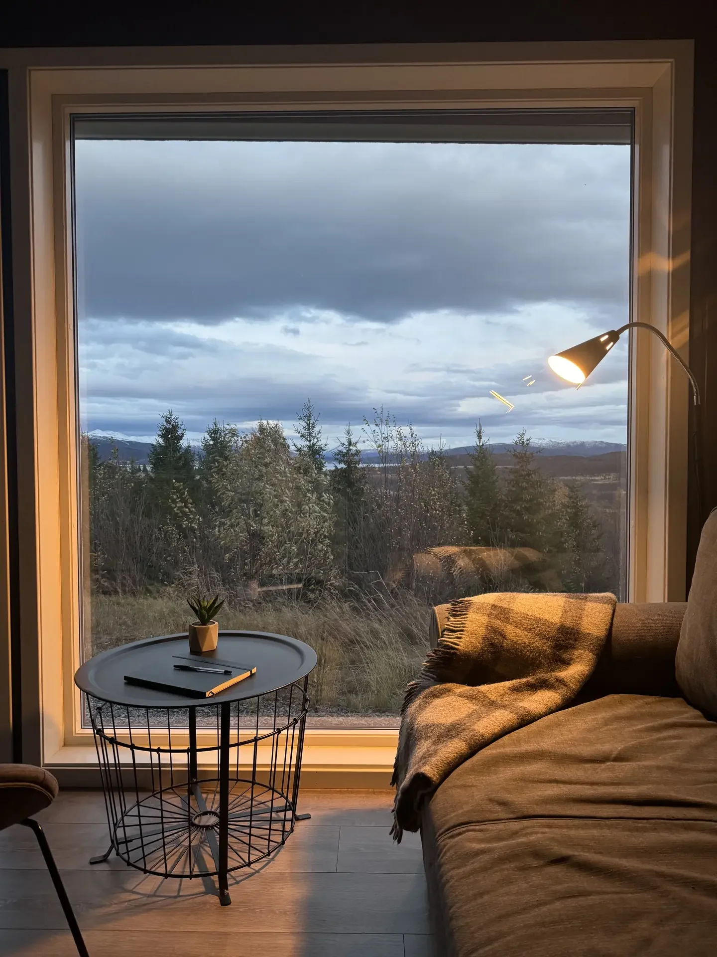 A warm cabin reading nook at dusk: a soft lamp, a wool throw and a notebook on a side table, framed by a picture window over the hills.