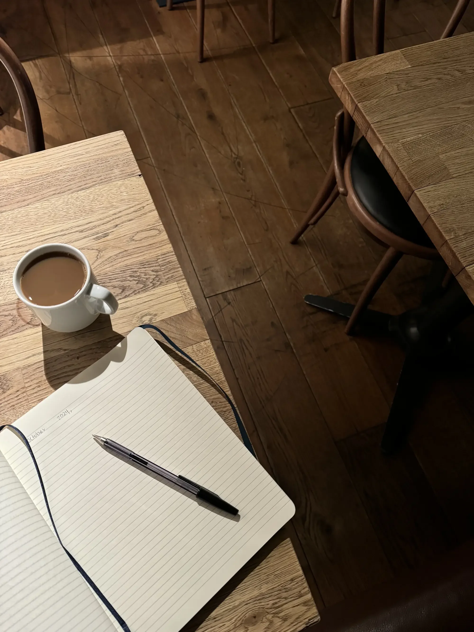 A cup of coffee and an open lined journal with a pen resting on it, on a warm wooden café table