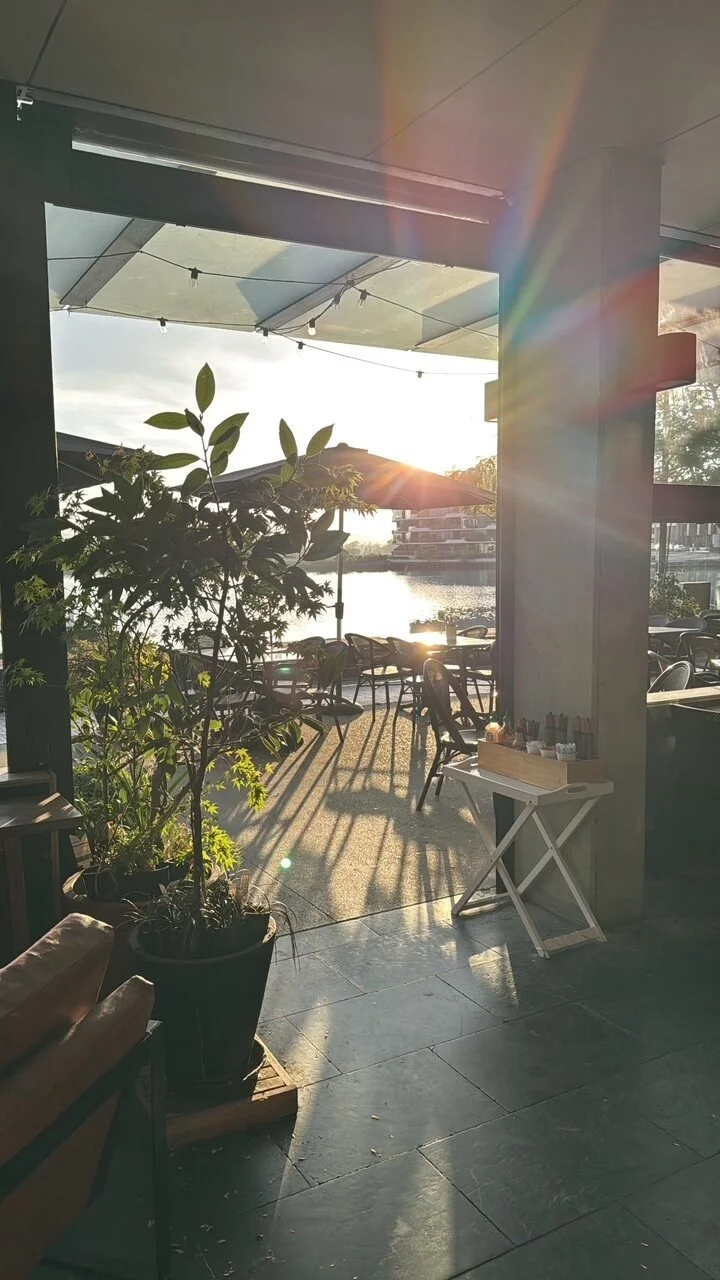 Golden-hour sunlight streaming through an open riverside café, with a potted plant and outdoor seating overlooking the water and a soft rainbow flare across the frame