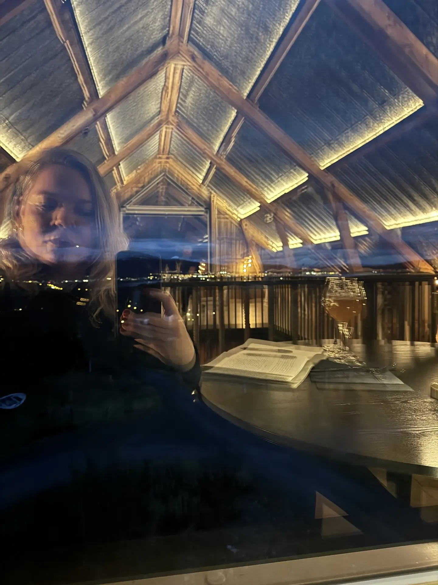 Paige reflected in the glass of a timber-vaulted lodge at dusk, phone in hand, an open book and the lights of a town below through the window.