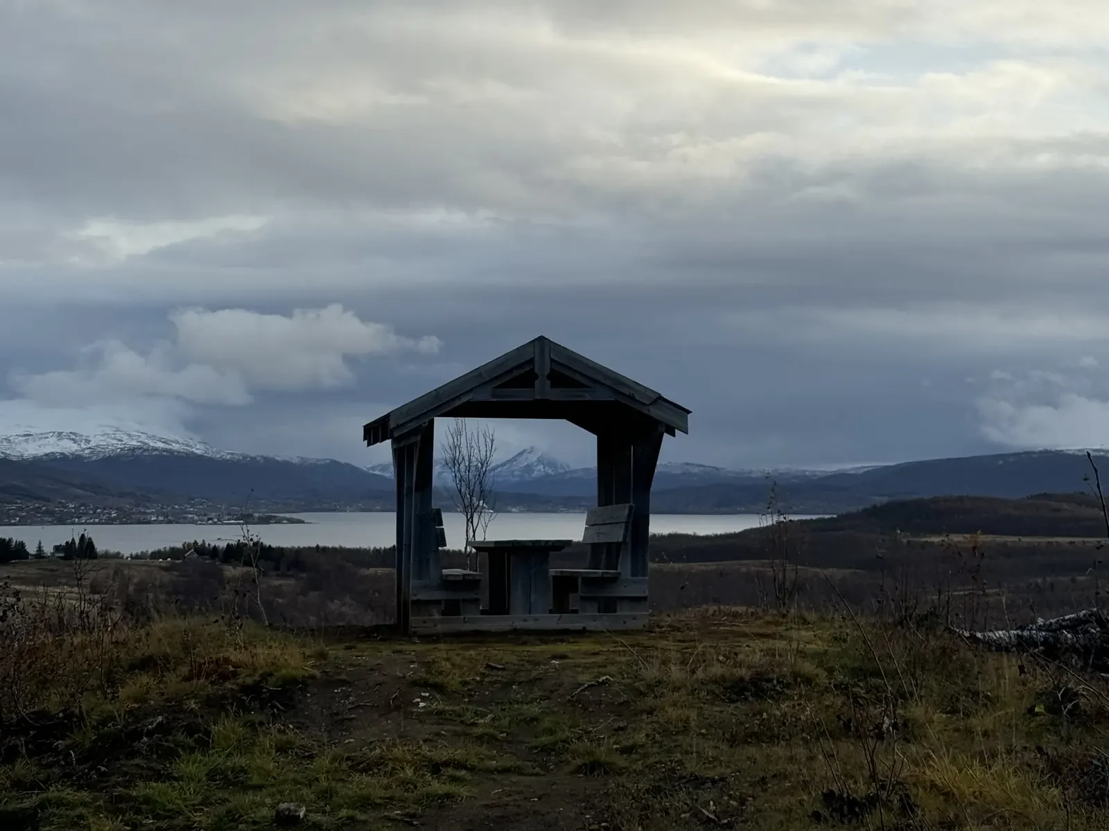 A simple timber shelter with a bench on a grassy headland, looking out over a calm Norwegian fjord and snow-dusted mountains under a soft, brooding sky.