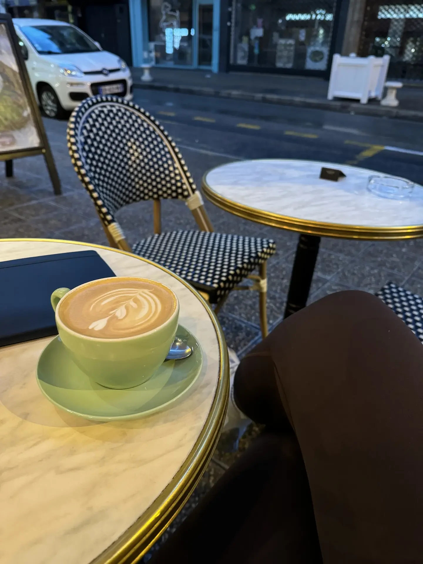 A latte in a sage-green cup on a marble bistro table outside a European street café, with a classic black-and-white rattan chair.