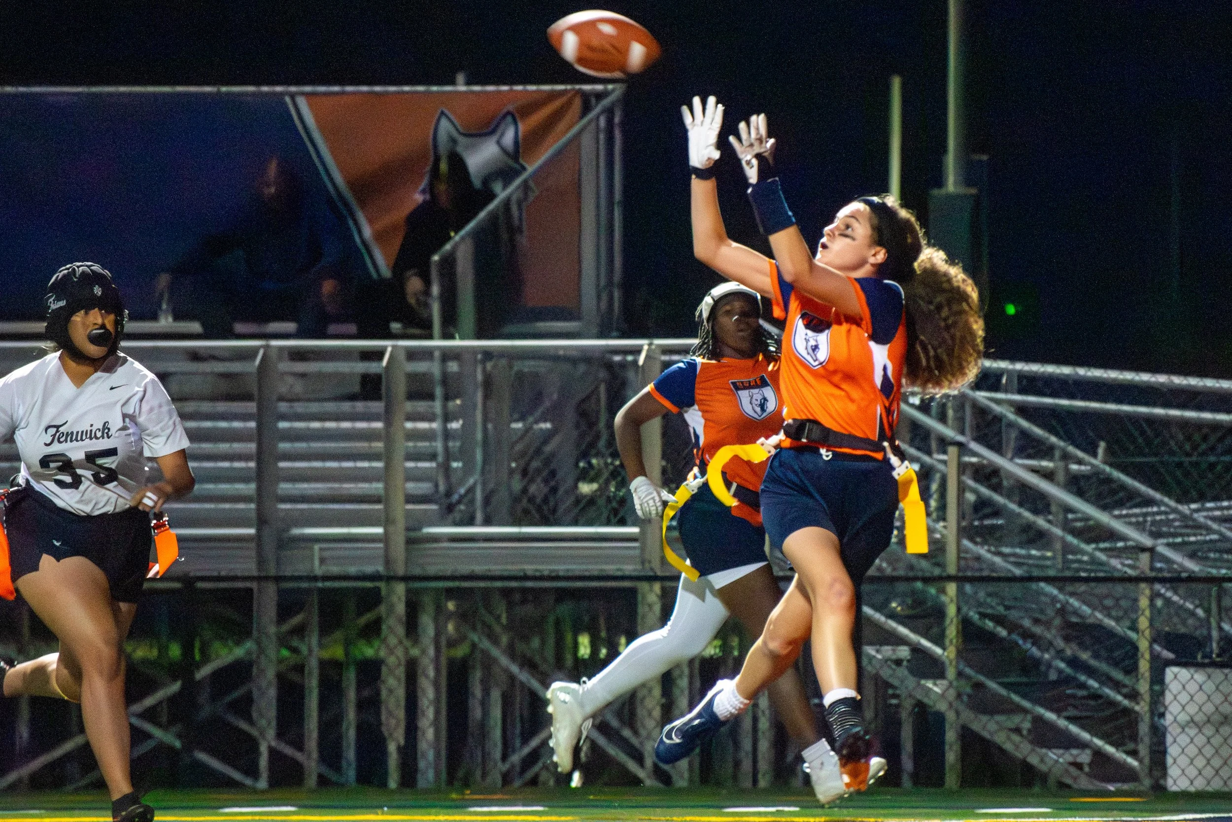 Female flag football players in orange and blue uniforms attempting to catch a football with one player jumping and reaching up during a game at night.