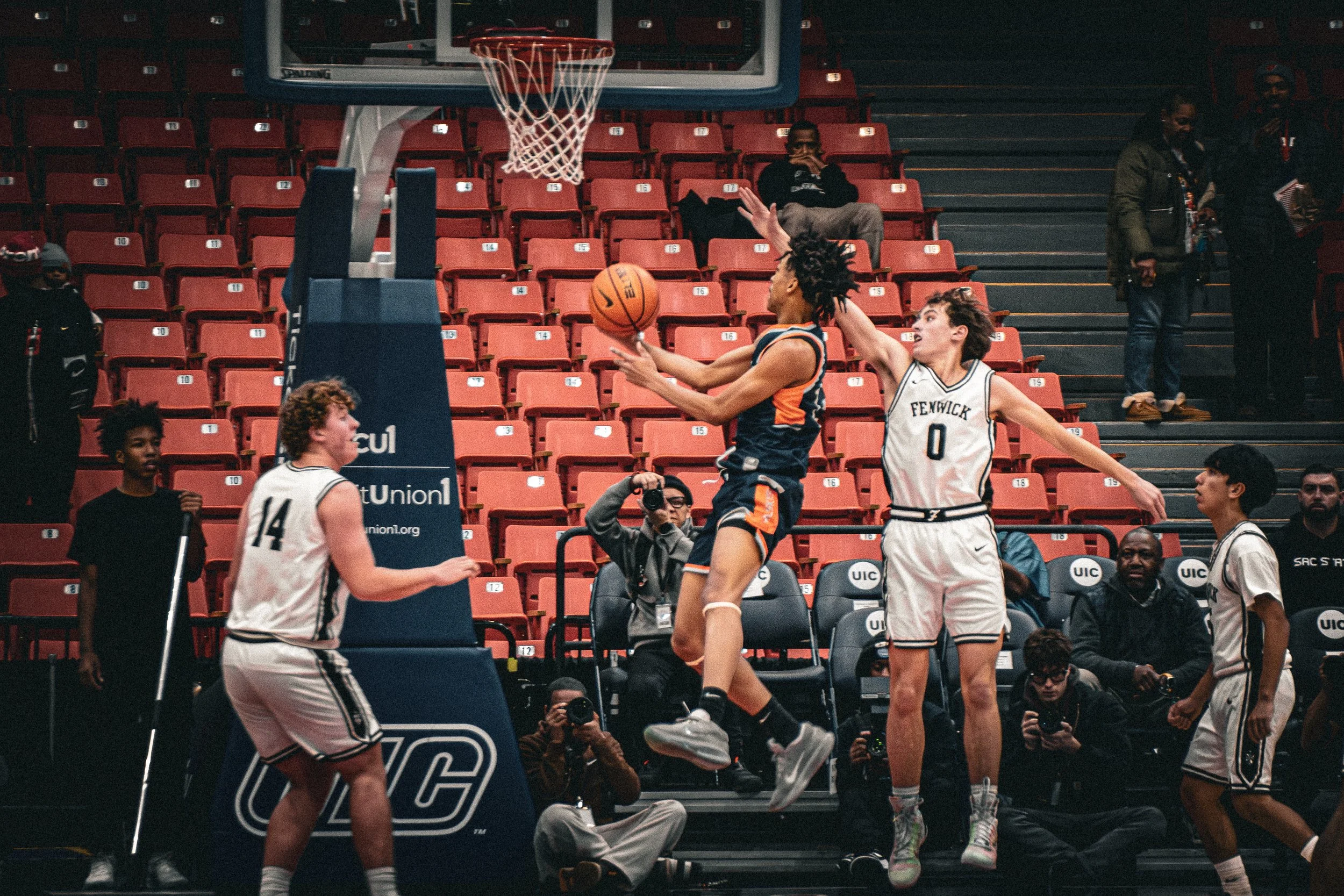 A basketball game with players from Fenwick and an opposing team. One player is jumping to shoot the ball while another from the opposing team attempts to block the shot. Multiple spectators and photographers are watching on the sidelines. The scene 
