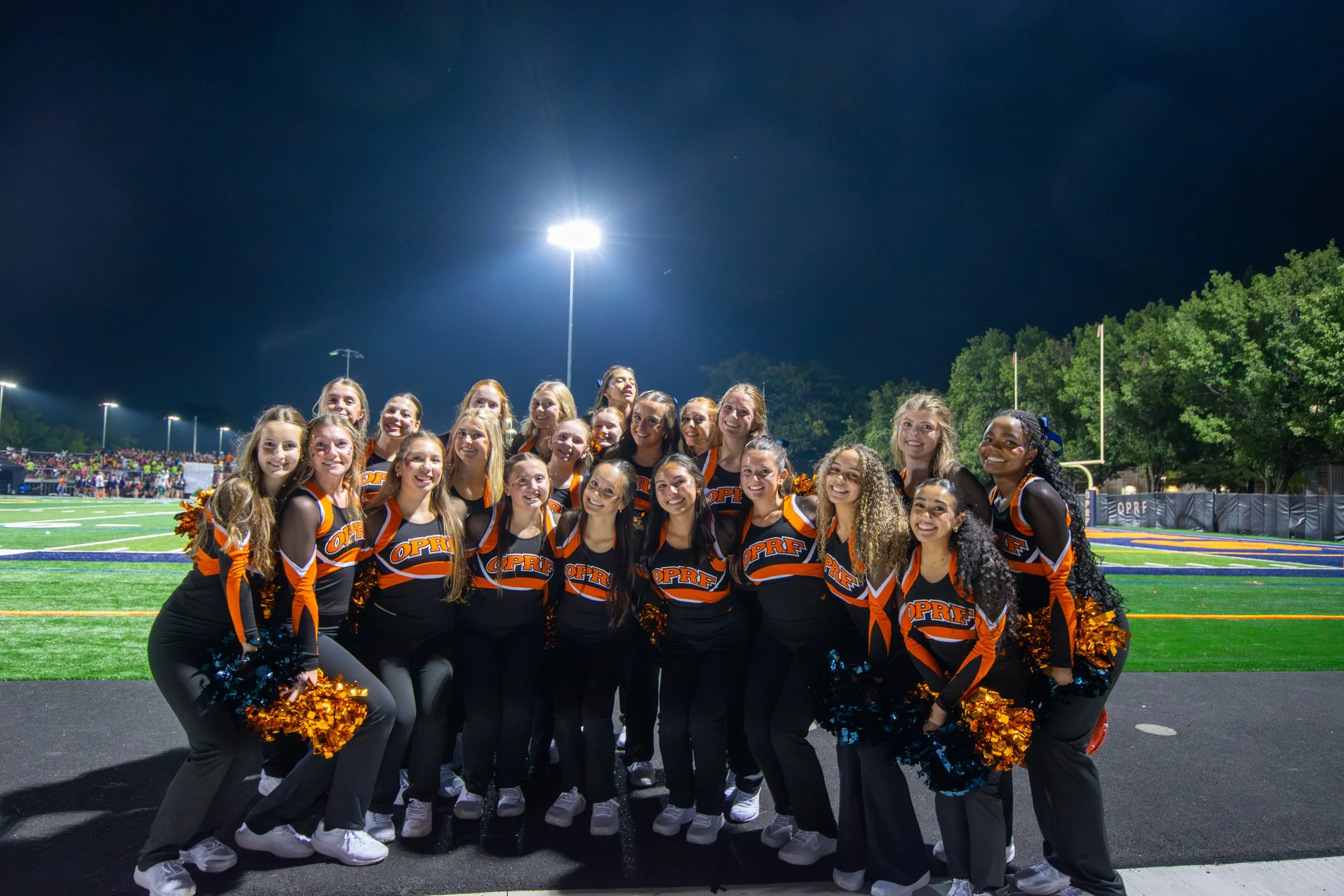 Cheerleaders in black and orange uniforms with 'OPRF' on the front, holding orange and black pom-poms, posing together on a football field at night under bright stadium lights, with a crowd in the background.