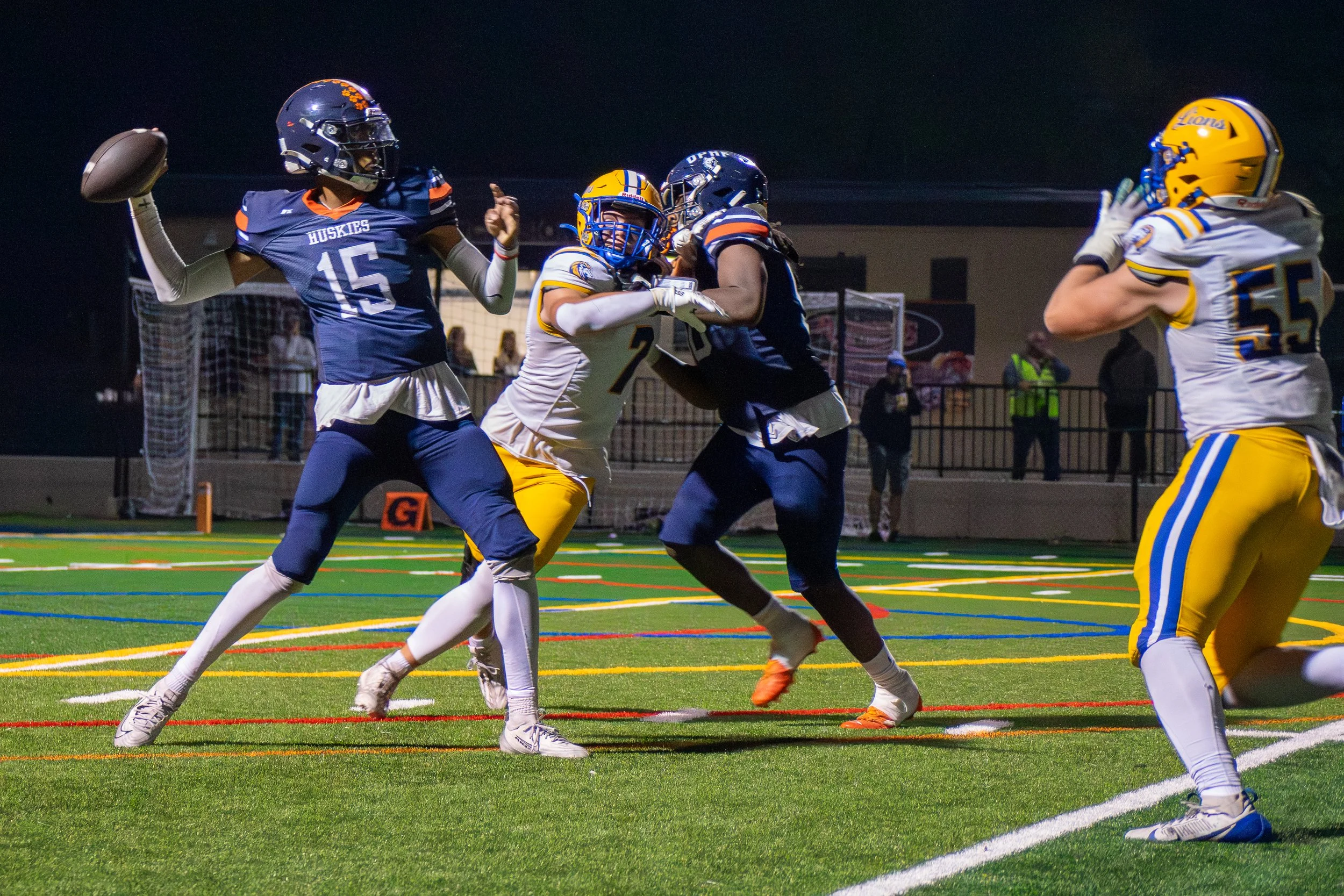Football players during a game, with one player in a blue jersey throwing a pass while teammates and opponents block and tackle on the field at night.