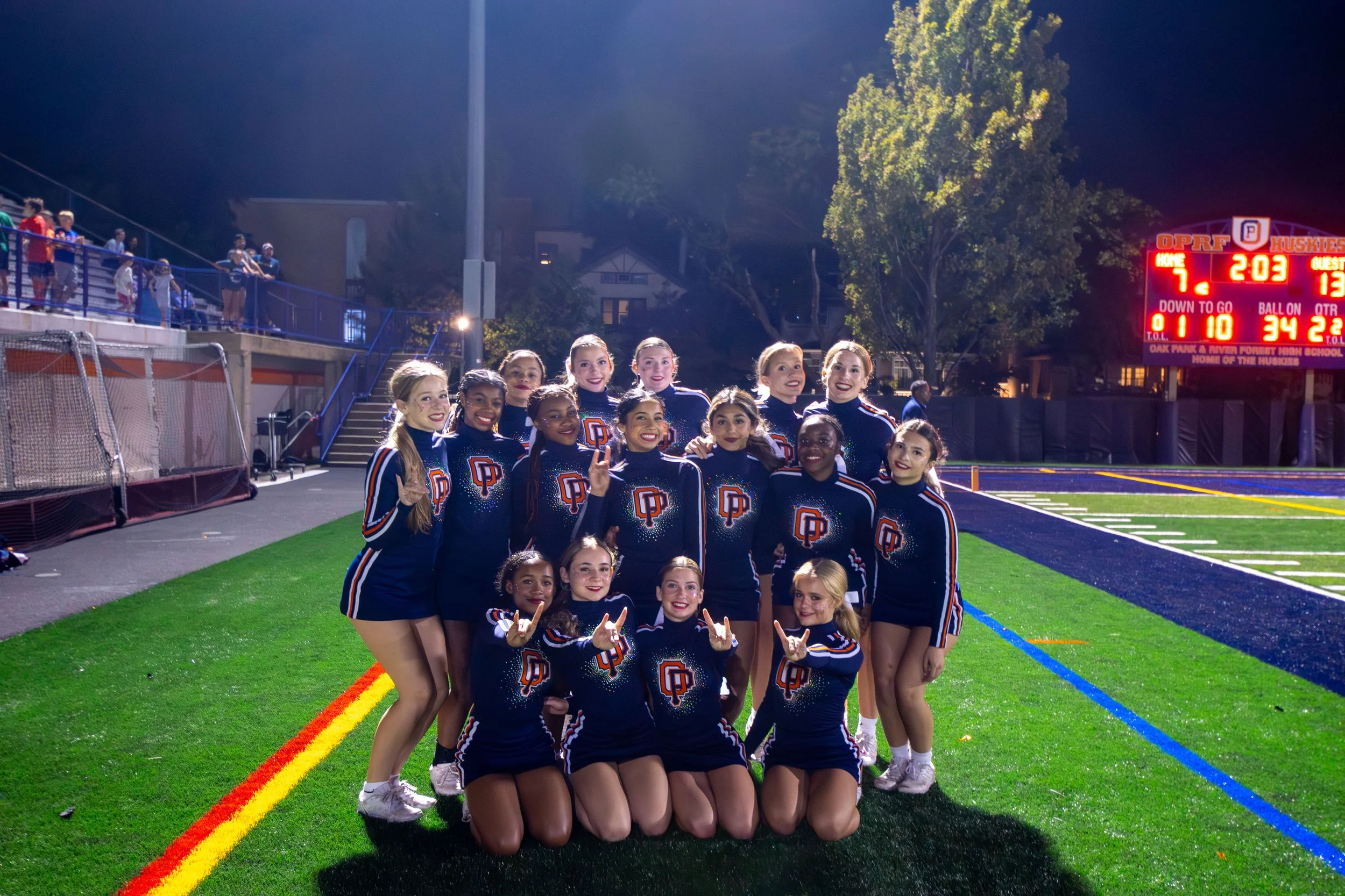 A group of cheerleaders in blue uniforms with 'DP' logos, posing on a football field at night, smiling and making hand gestures.