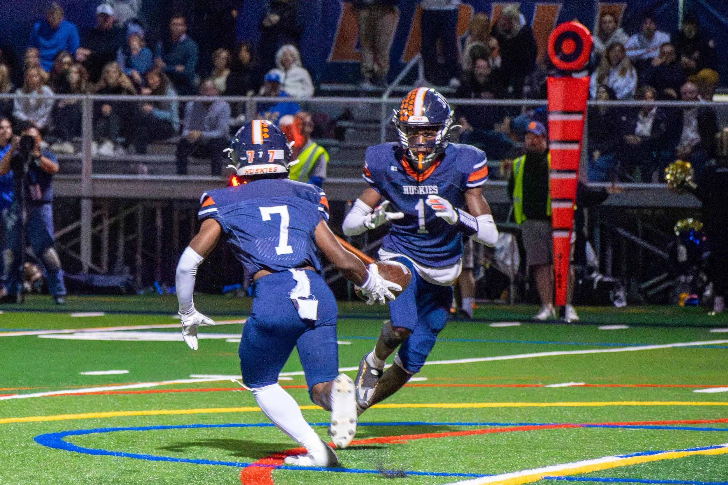 Two American football players from the Huskies team wearing blue uniforms, one with jersey number 7 and the other with 1, on a field during a game. One player is holding the football while the other is running towards him. The game is taking place at
